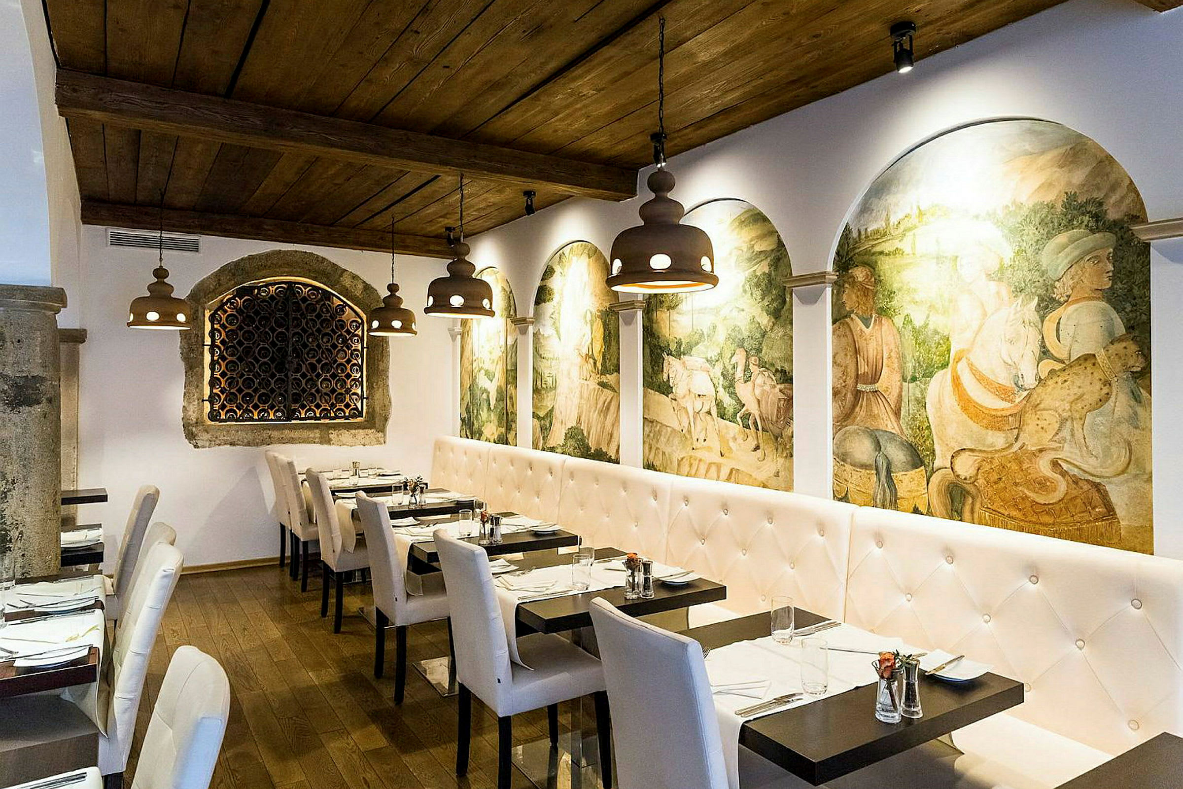 En restaurantinteriør med hvite polstrede sofaer og stoler, mørke trebord og en vegg dekorert med historiske freskomalerier. Taket er av tre, og belysningen består av hengende lamper. (AI-generert tekst)