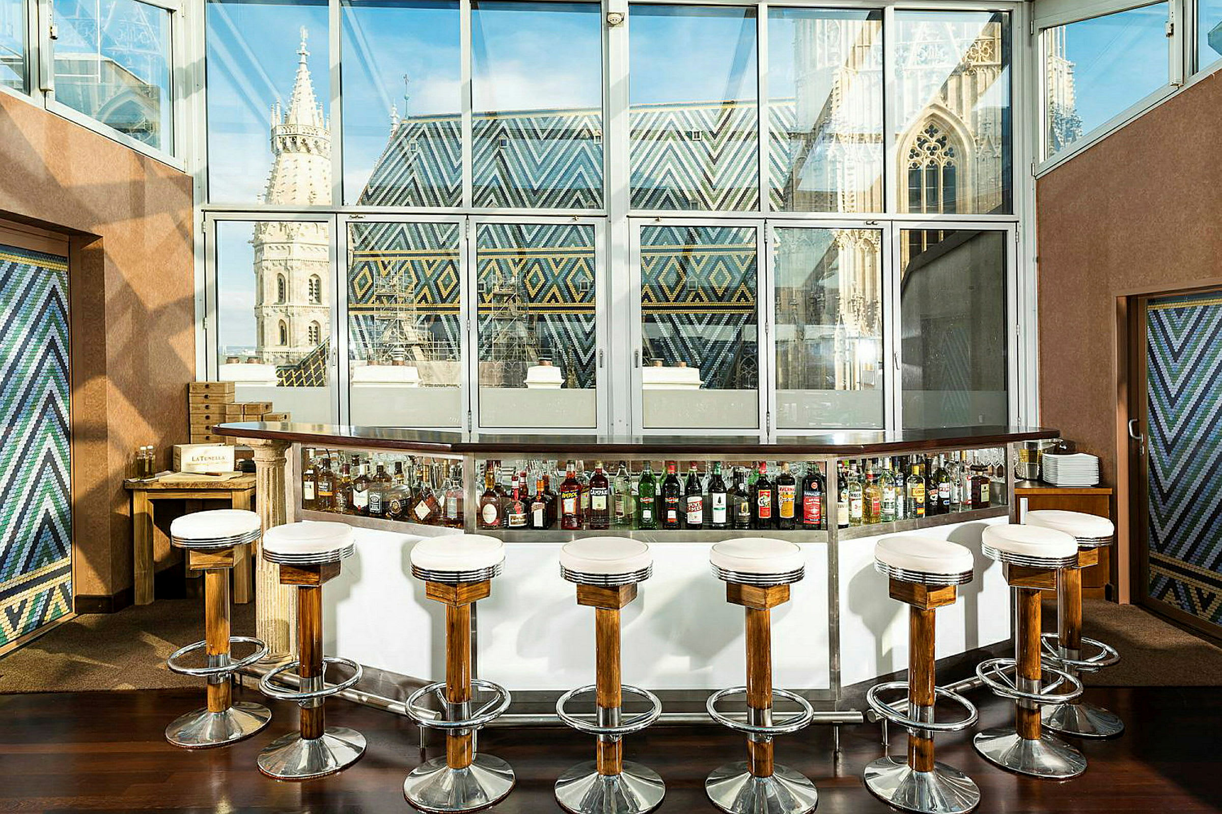 En cocktailbar med høye stoler foran en glassvegg med utsikt over en bygning med fargerike takdekorasjoner. (AI-generert tekst)