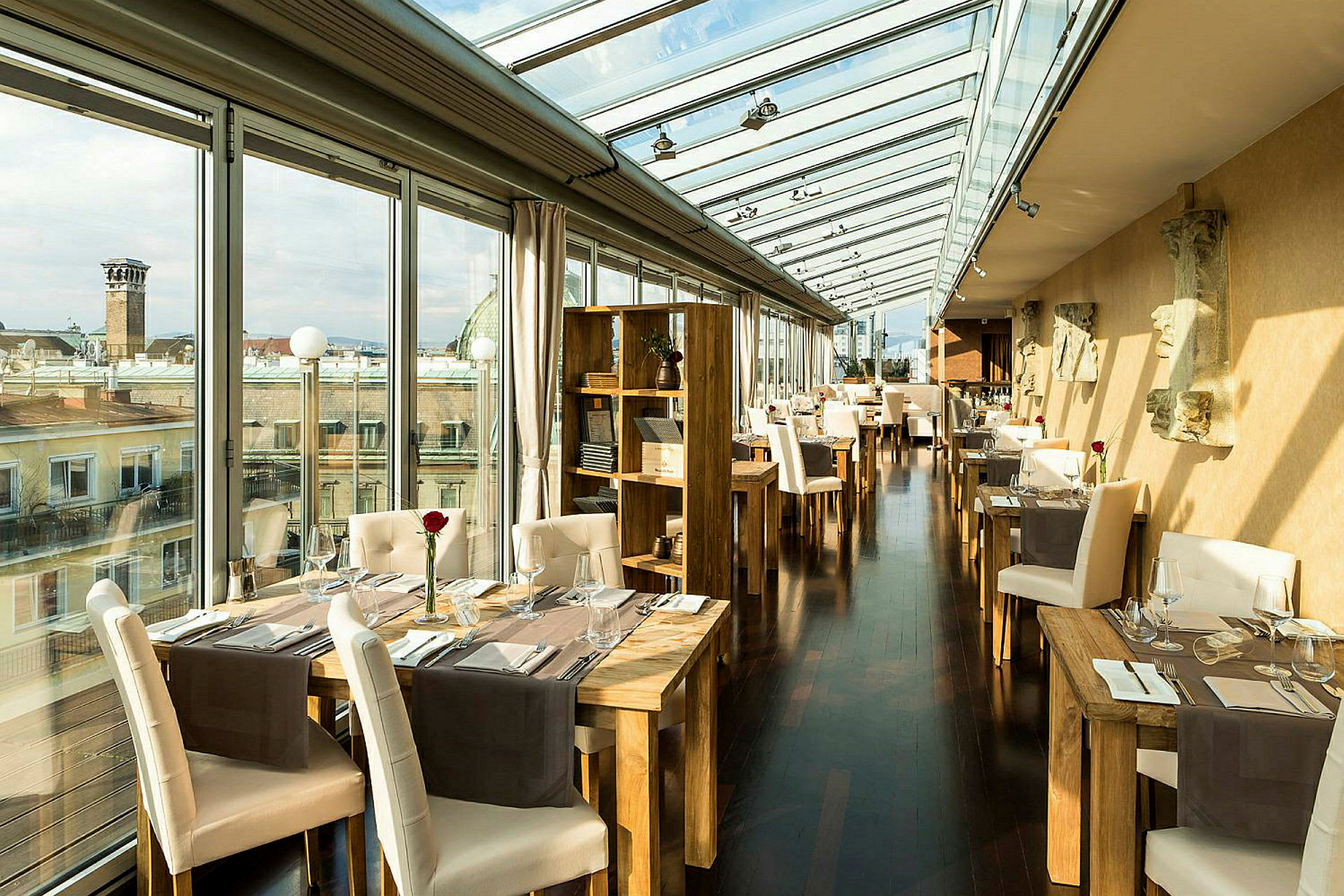 Et elegant restaurantområde med store glasspaneler som gir utsikt over bytakene. Bord og stoler er elegant satt opp med røde roser i glassvaser som borddekorasjoner. (AI-generert tekst)
