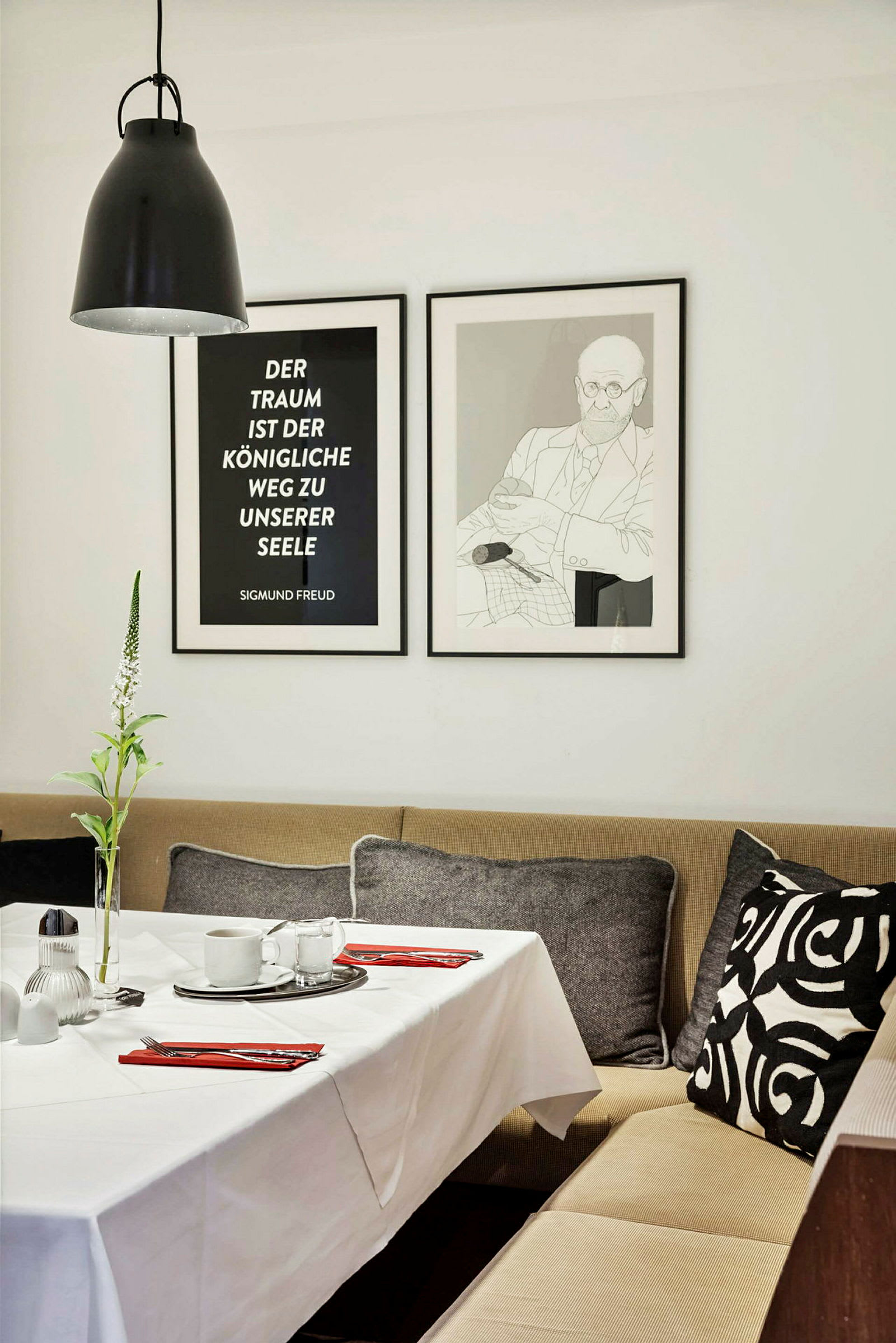 Et dekket spisebord med en hvit duk, røde servietter og porselen. I bakgrunnen er det en beige sofamøbel med pyntputer og to innrammede bilder på veggen. (AI-generert tekst)