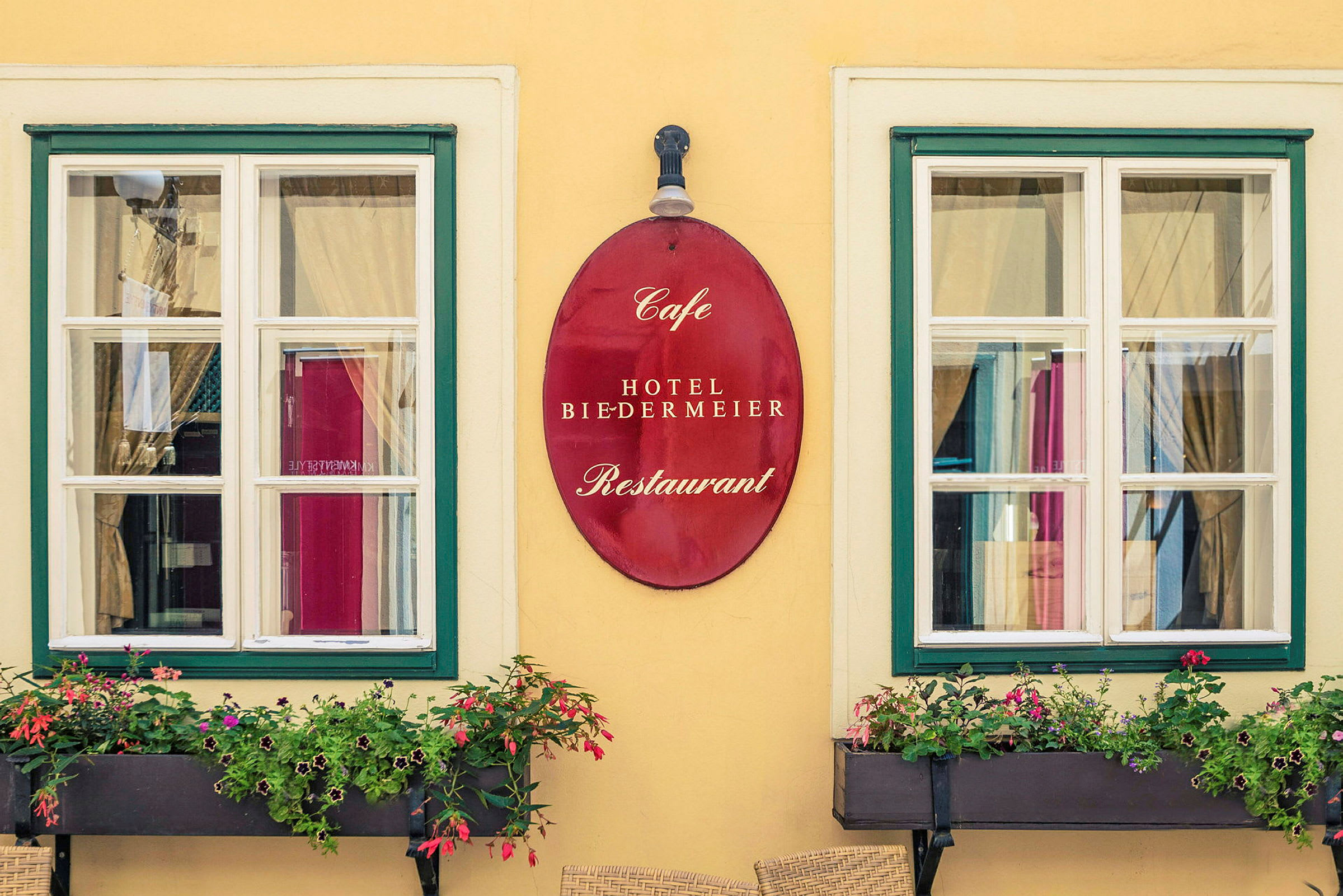 To vinduer med hvite karmer og grønne rammer, mellom dem henger et rødt skilt med teksten "Cafe Hotel Biedermeier Restaurant". Foran vinduene er blomsterkasser med blomstrende planter. (AI-generert tekst)