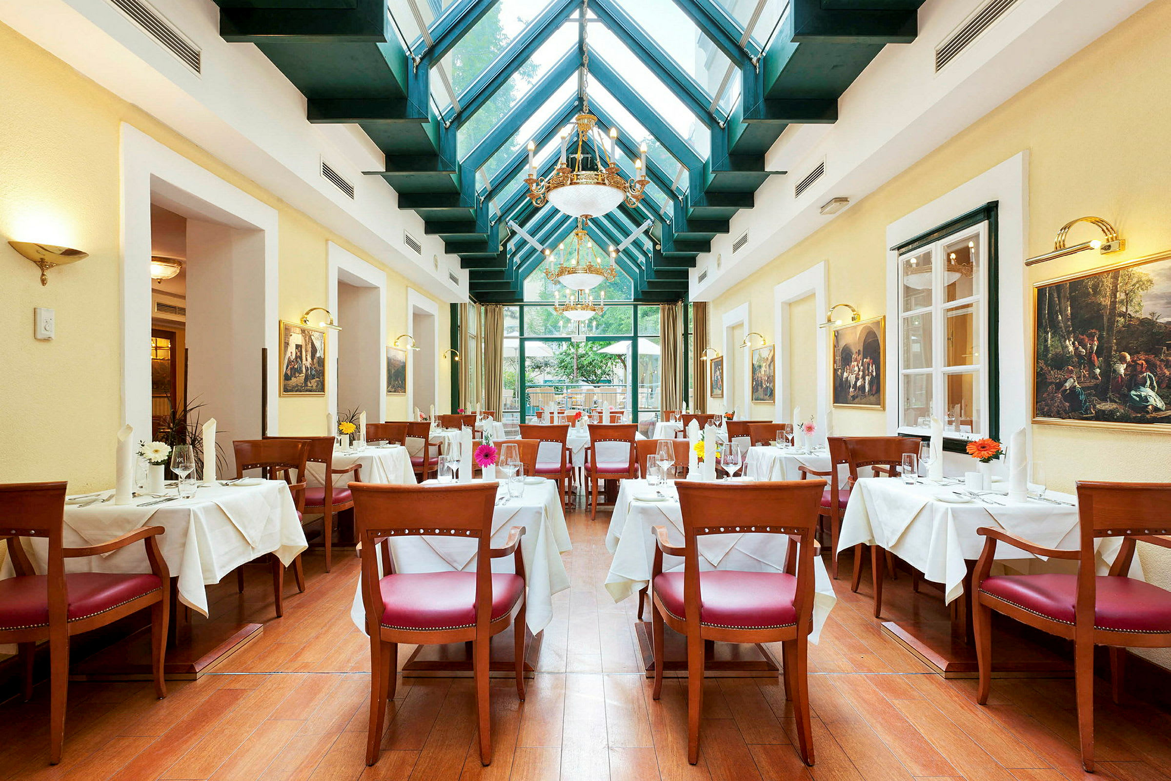 En elegant restaurant med høyt tak og glasstak. Bordene er dekket med hvite duker, og stolene har røde seteputer. Veggene er dekorert med malerier. (AI-generert tekst)