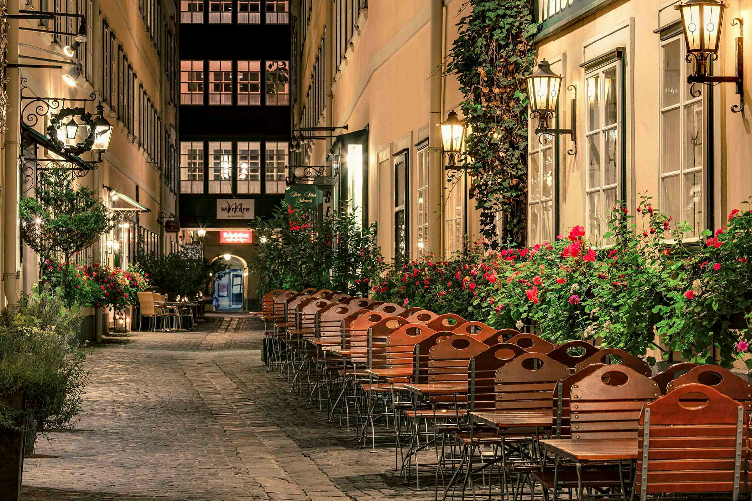 En pittoresk gate med stoler og bord utenfor restauranter, omgitt av blomsterbusker og lamper. (AI-generert tekst)