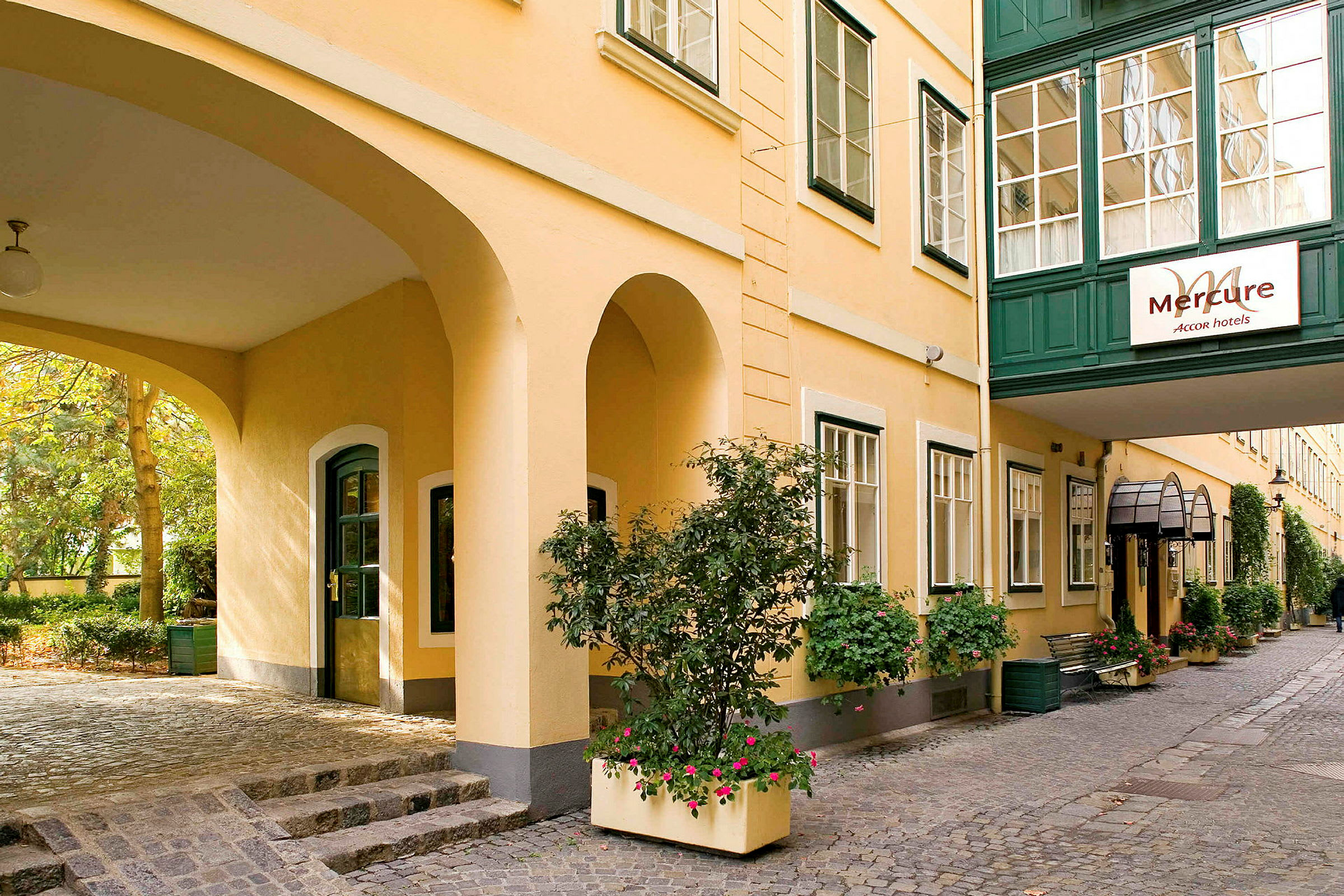 En gårdsplass ved hotell Mercure med gule vegger og grønne vindusrammer, dekorert med blomster og grønn vegetasjon. (AI-generert tekst)