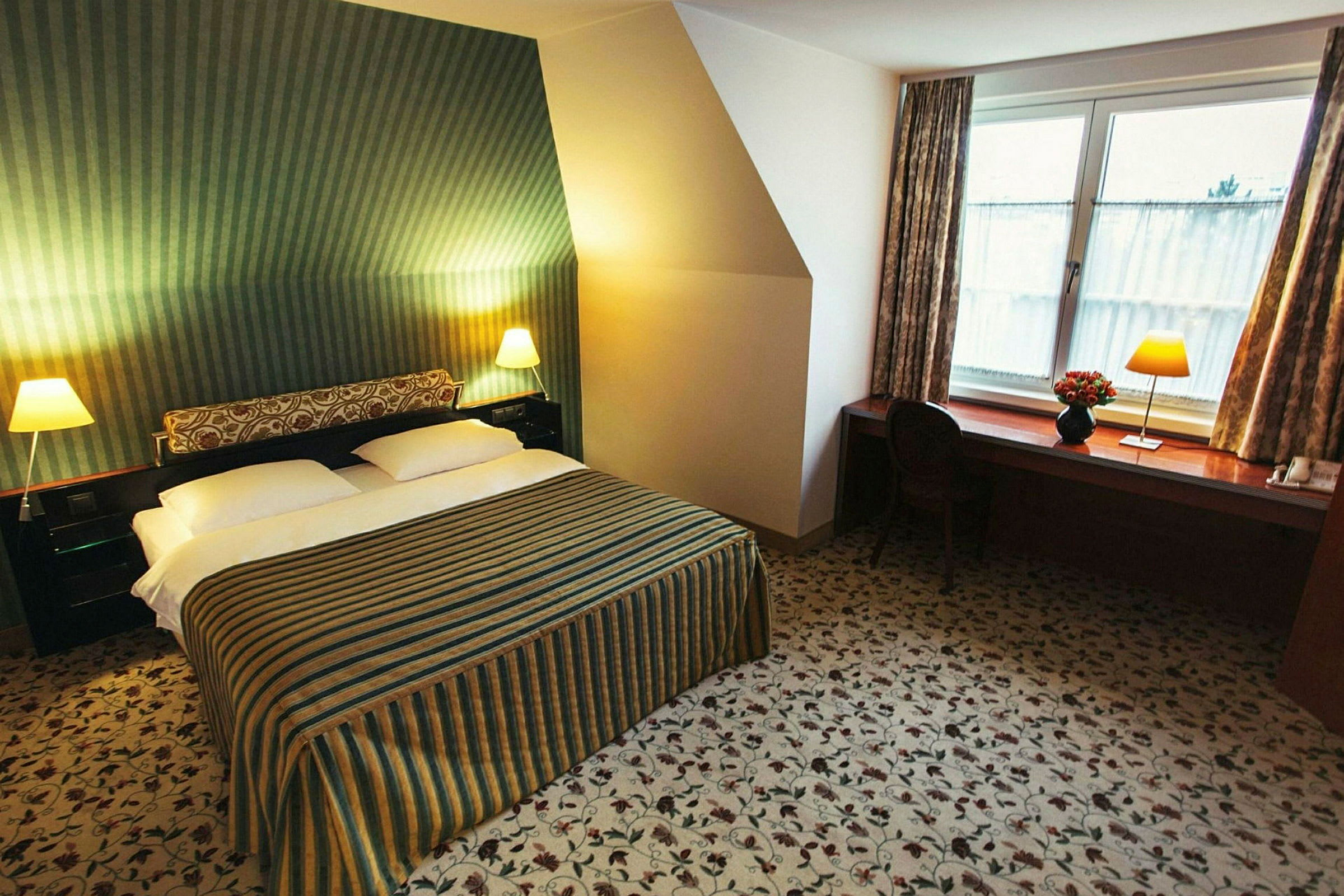 Et hotellrom med en dobbeltseng, stripete sengeteppe, to nattbord med lamper og en arbeidsbenk med stol ved et vindu. (AI-generert tekst)