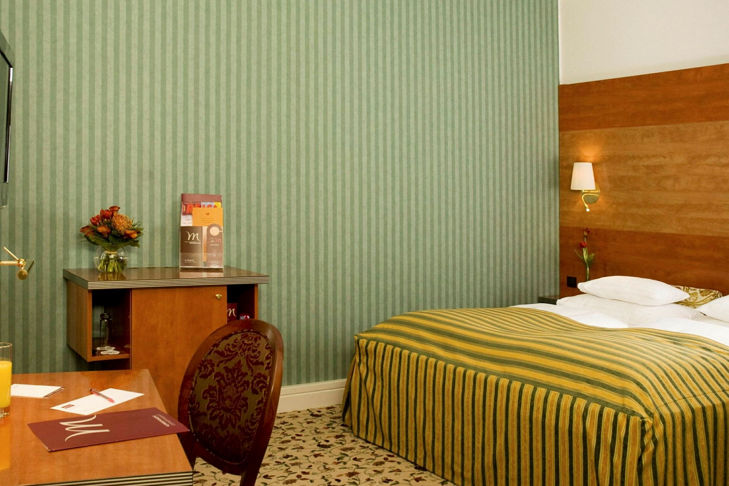 Et hotellrom med en seng med stripete gulgrønt sengeteppe, et skrivebord og en stol. Det er en blomsterkrukke og informasjonsmateriale på skrivebordet. (AI-generert tekst)