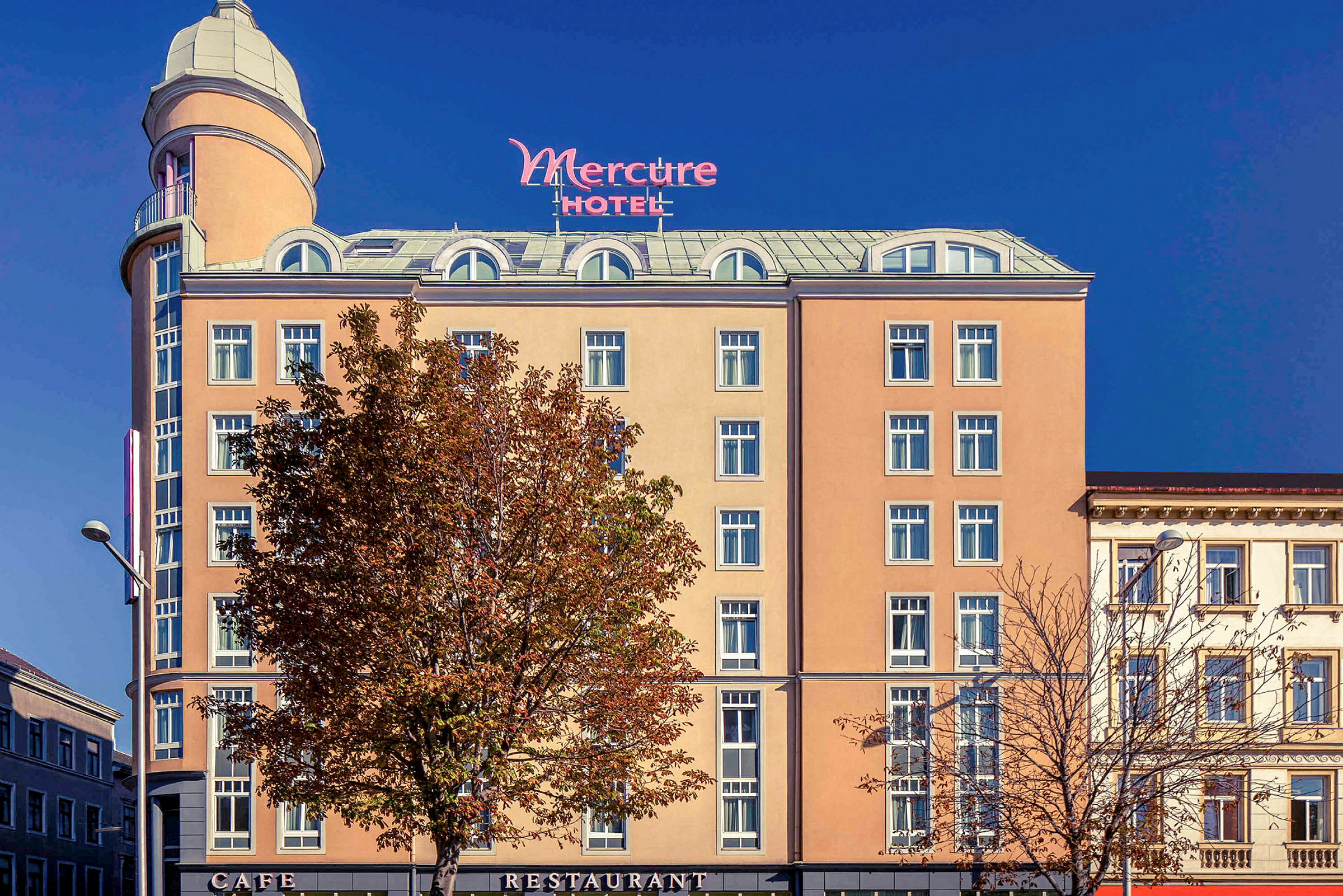 Eksteriøret av et hotell med navnet Mercure, med en beige fasade, mange vinduer og et høyt tårn på toppen. (AI-generert tekst)