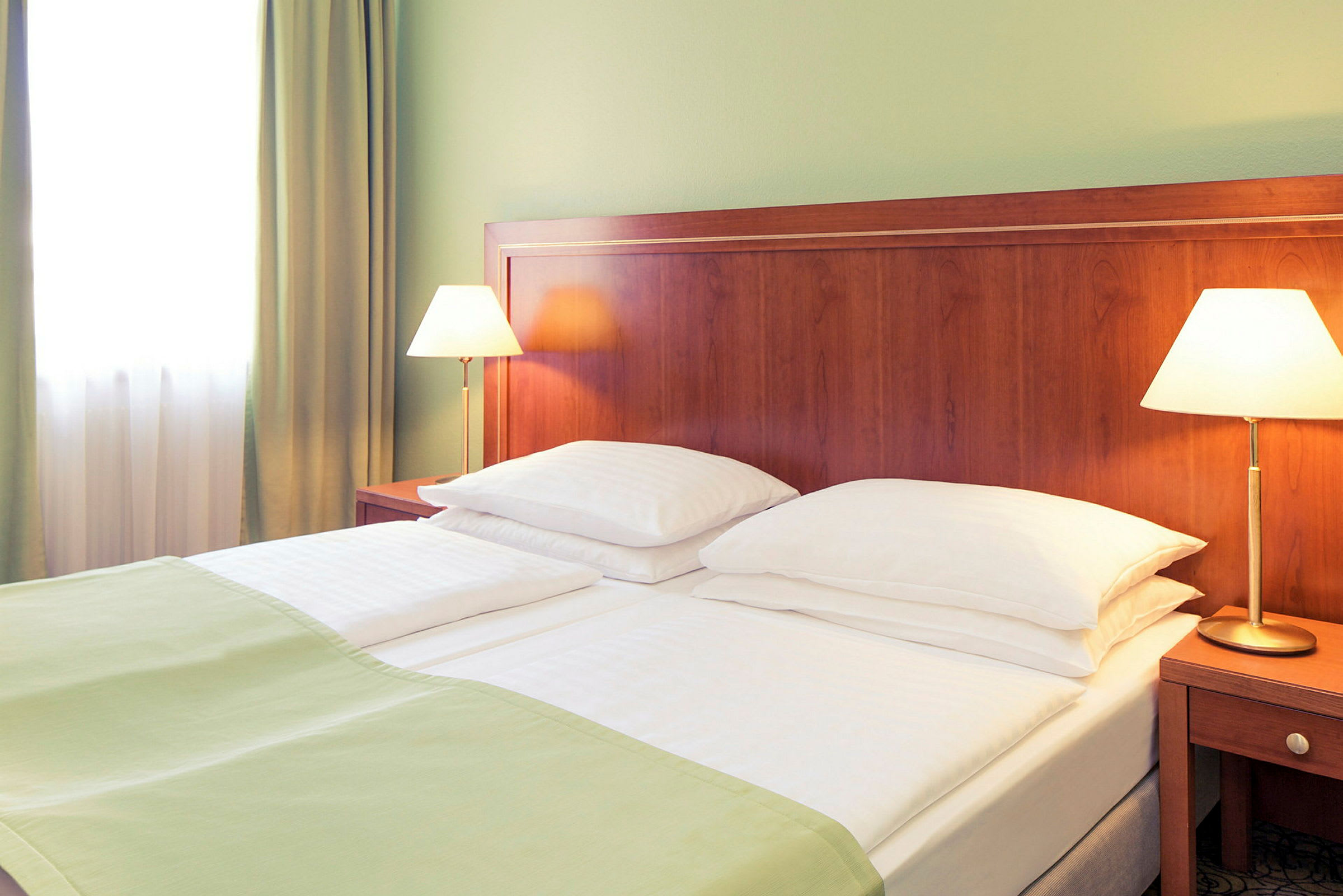 Et hotellrom med en dobbeltseng som har hvite sengeklær og grønne tepper. To sengelamper står på trebord ved sengen. (AI-generert tekst)