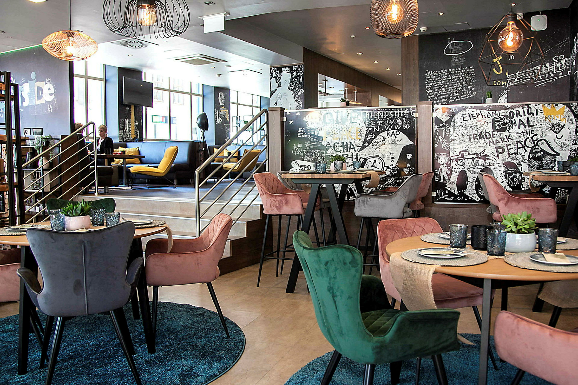 En moderne interiør av en restaurant med fargerike stoler rundt bord som har dekketallerkener og glass. Veggene er dekorert med kunstneriske mønstre og tekster. (AI-generert tekst)