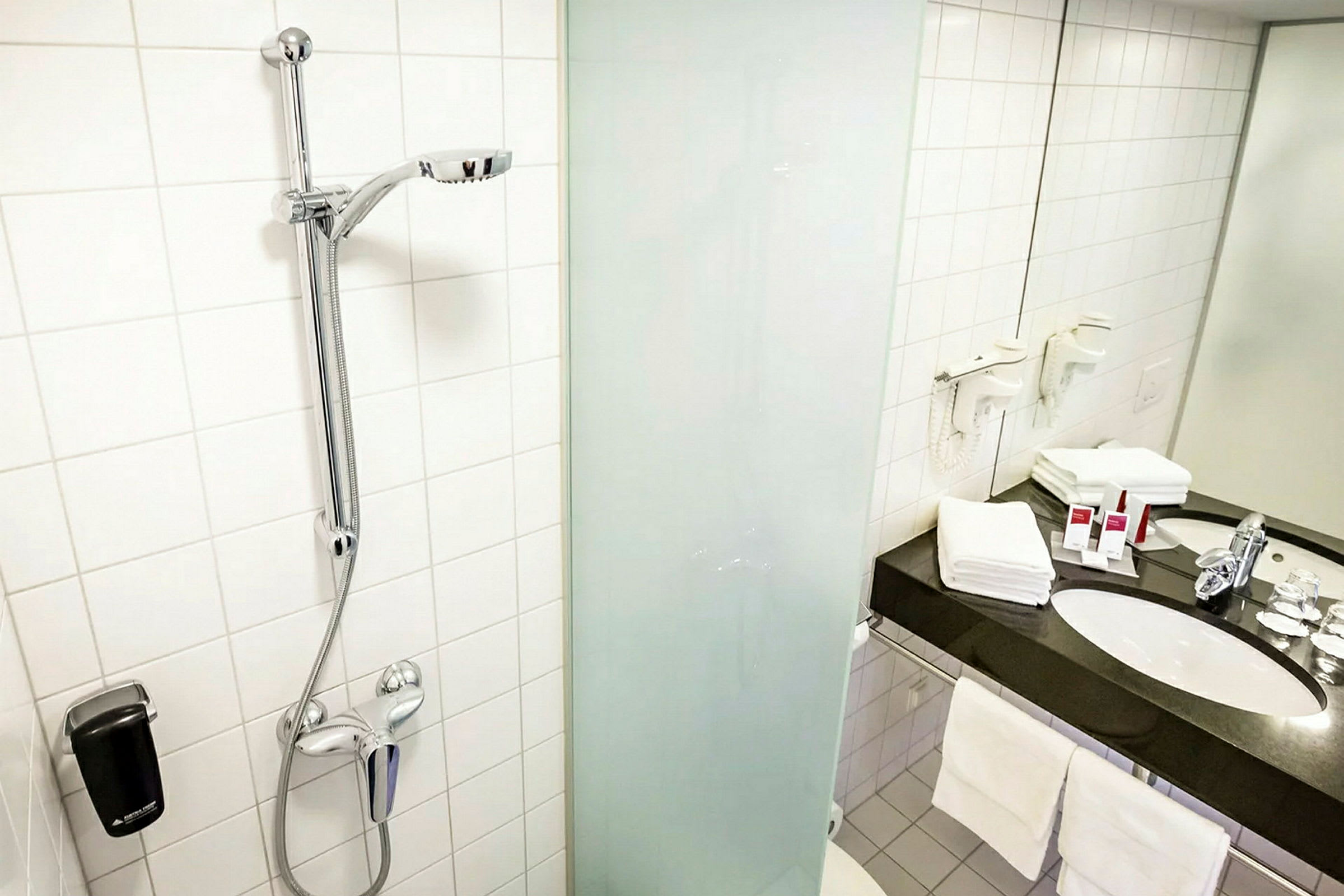 Et baderom med en dusj med glassvegg og en hånddusj. En benkeplate med servant, speil og håndklær er også synlige. (AI-generert tekst)