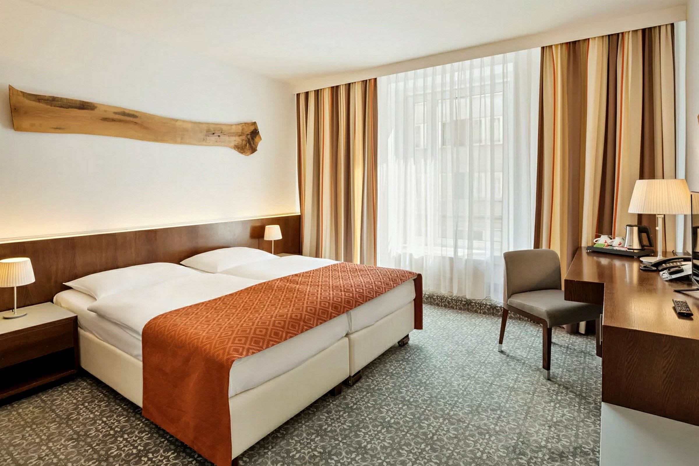 Et hotellrom med en dobbeltseng som har et oransje sengeteppe og to hvite puter. Det er nattbord med lamper på begge sider og et stort vindu med gardiner i bakgrunnen. Et skrivebord med en stol og diverse gjenstander står til høyre. (AI-generert tekst)