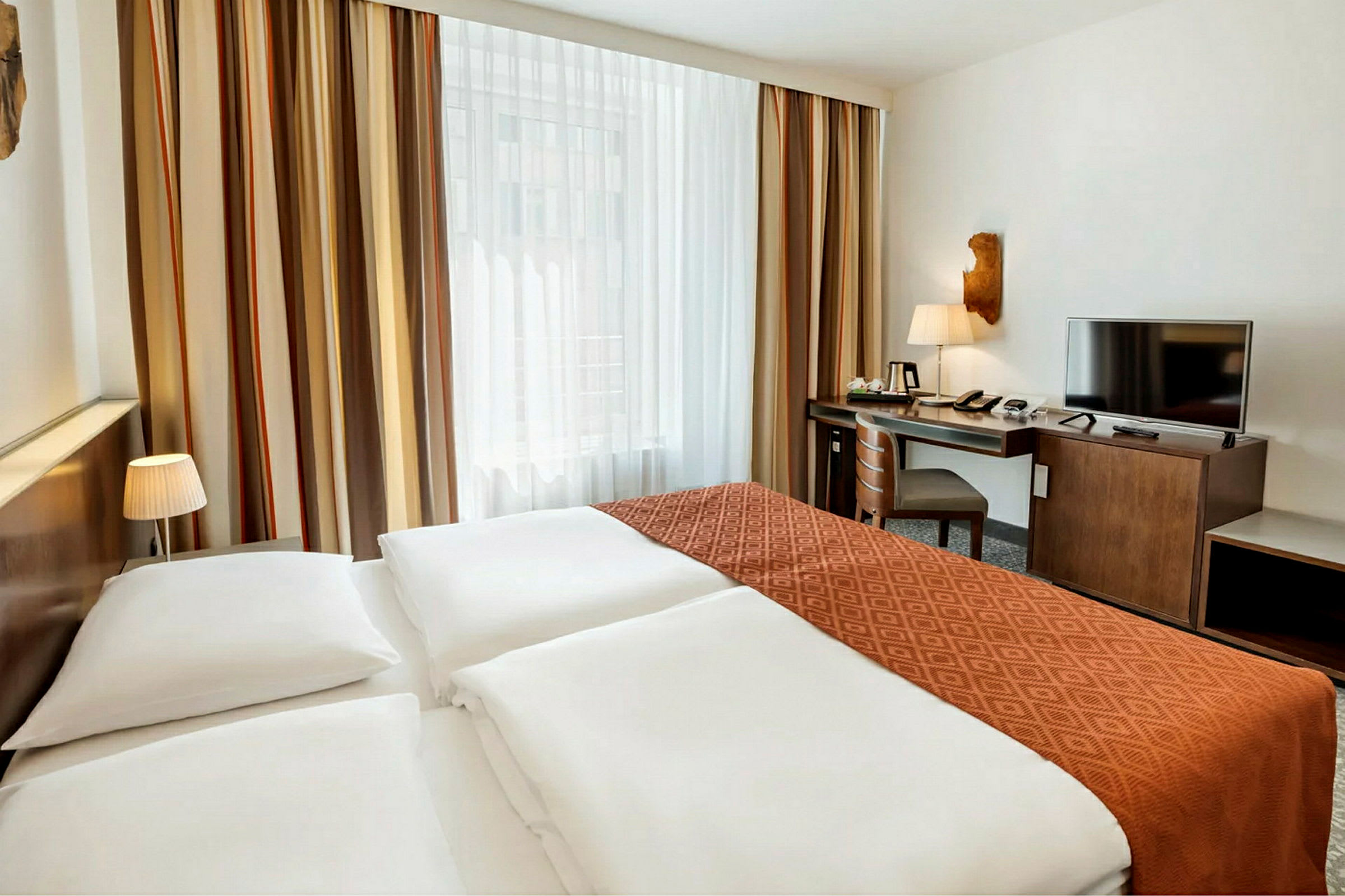 Hotellrom med en bred seng, oransje sengeteppe, vindu med gardiner samt et skrivebord med stol og TV. (AI-generert tekst)