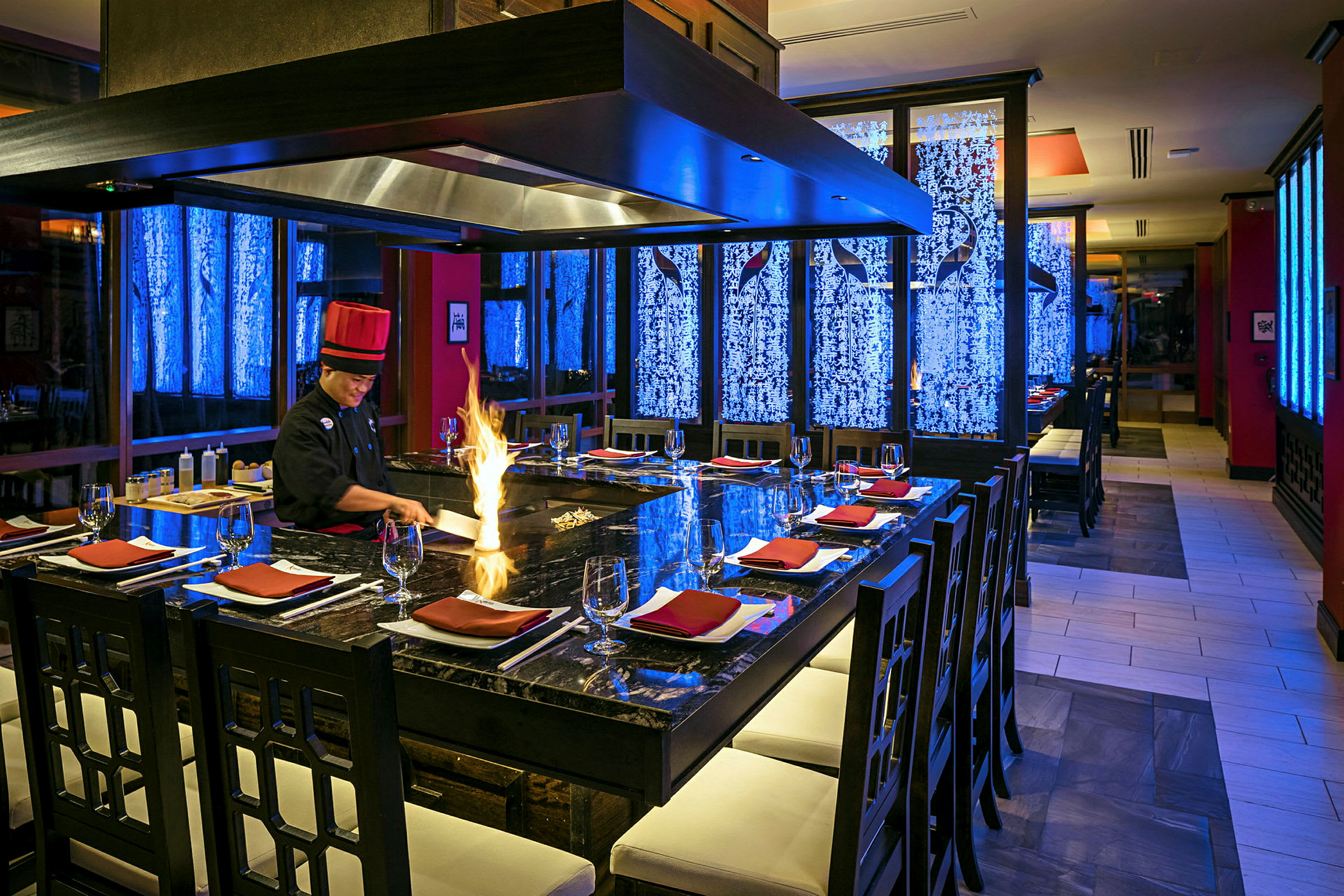 Restaurant med teppanyaki-kokk ved en stor matlagingsstasjon, omgitt av stoler og borddekning. Flammer sees på grillen. (AI-generert tekst)
