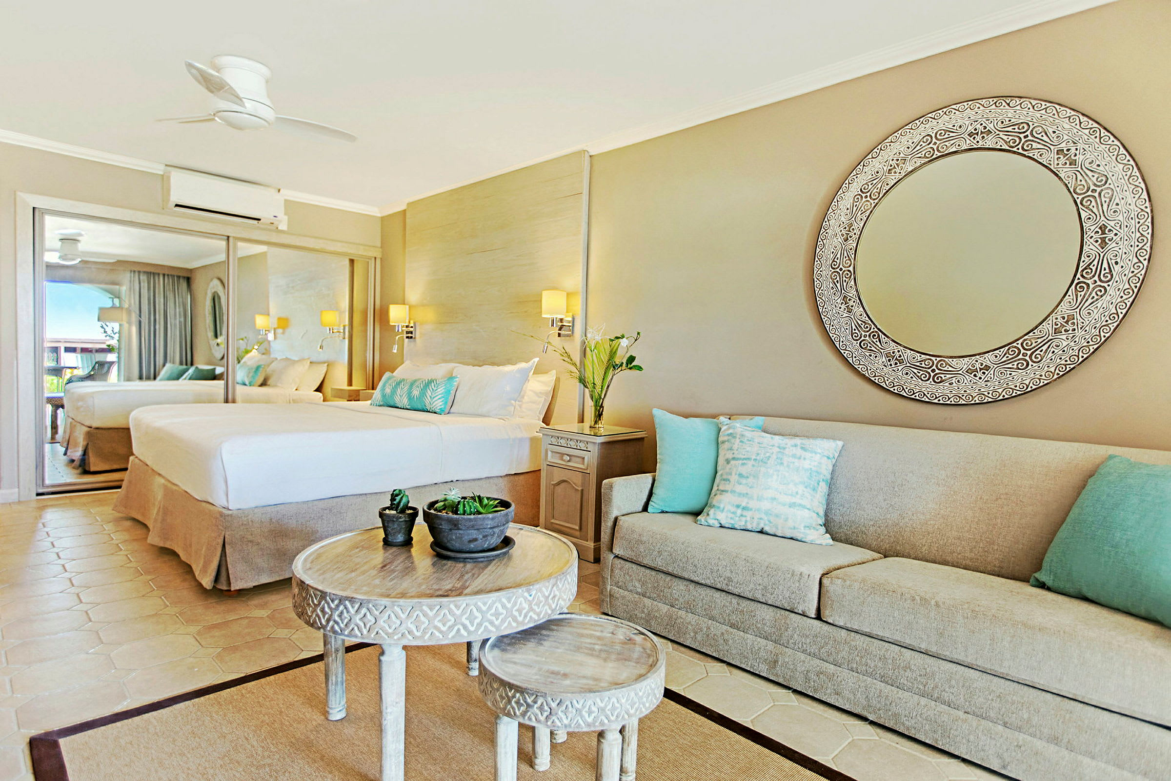 Et elegant hotellrom med et stort speil, en bred seng og en beige sofa med dekorative puter. Et rundt bord med mindre bord og plantepynt i midten. (AI-generert tekst)