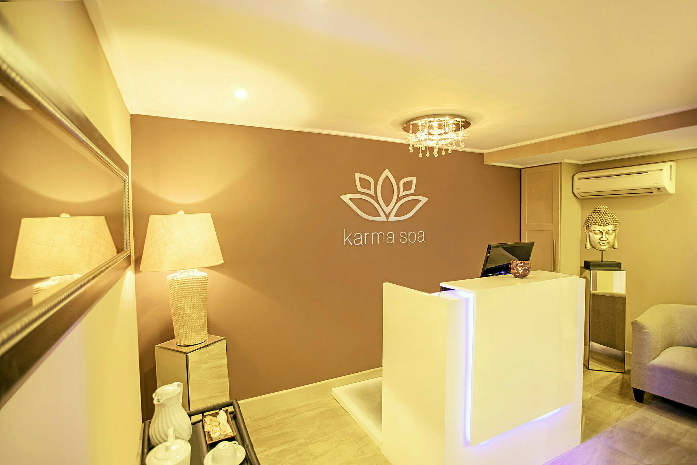 En resepsjon i et spa-anlegg med en moderne og elegant design. En hvit resepsjonsdisk står foran en vegg med logo, og en lampe lyser opp rommet. (AI-generert tekst)