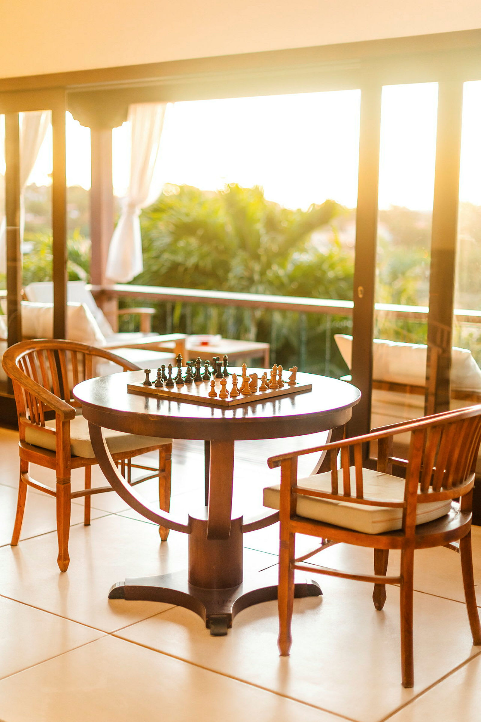 Et sjakkbrett står på et bord omgitt av stoler på en terrasse med utsikt over grøntområder. (AI-generert tekst)