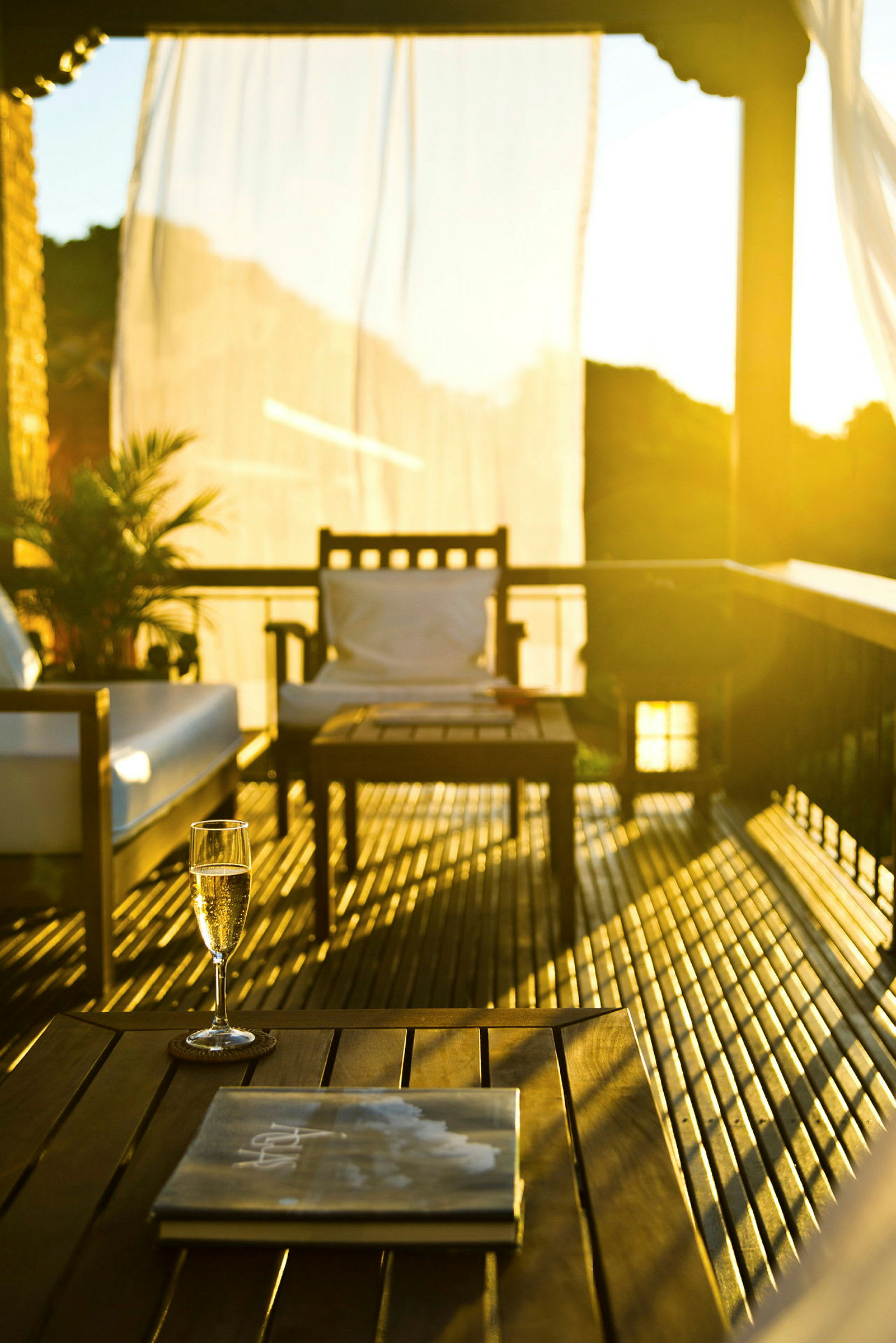 En veranda med møbler inkludert et bord, en stol og en sofa, opplyst av gyllent sollys. På bordet står et glass og en bok. (AI-generert tekst)