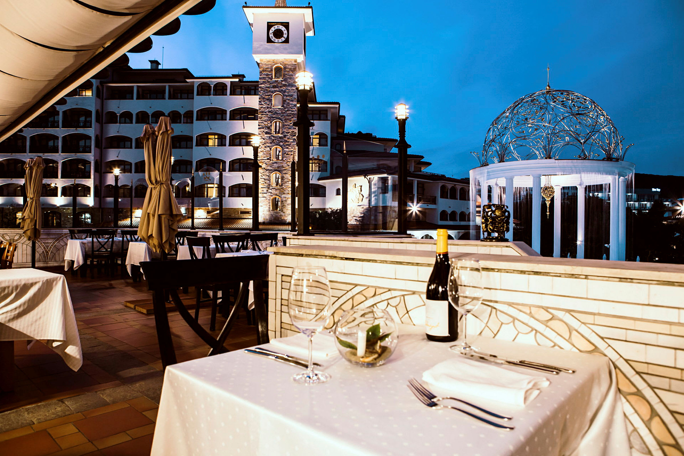 En uterestaurant med dekkede bord og utsikt over en bygning med et klokketårn og en opplyst paviljong. (AI-generert tekst)
