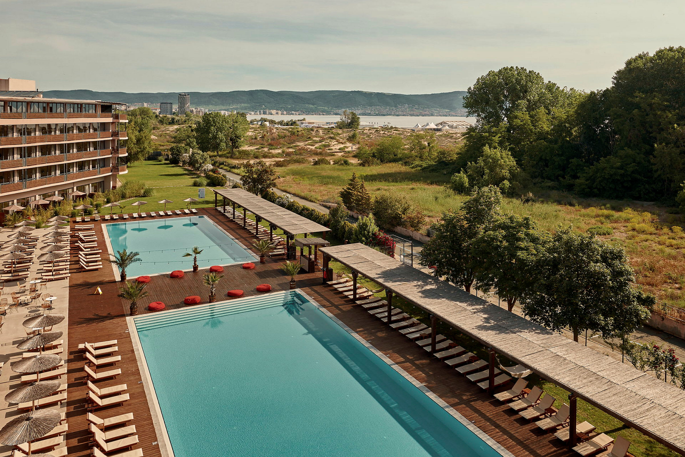 Stor basseng med solsenger og parasoller ved siden av et hotell, med grøntområder og landskap i bakgrunnen. (AI-generert tekst)