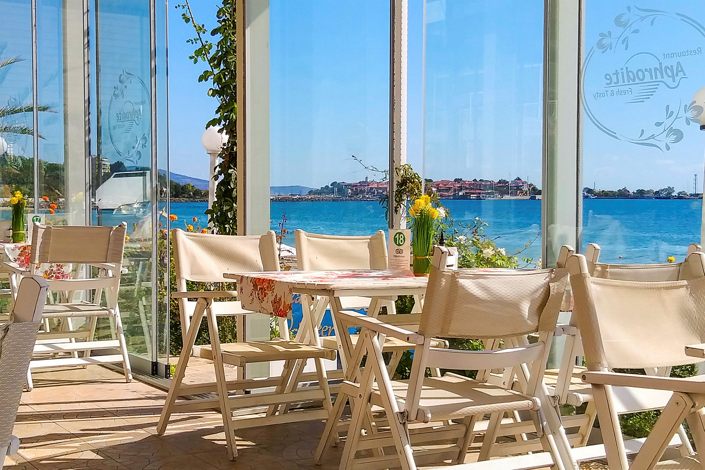 Utsikt fra en restaurant med hvite stoler og bord, med glassvegger som gir utsyn over havet og en kystby. (AI-generert tekst)