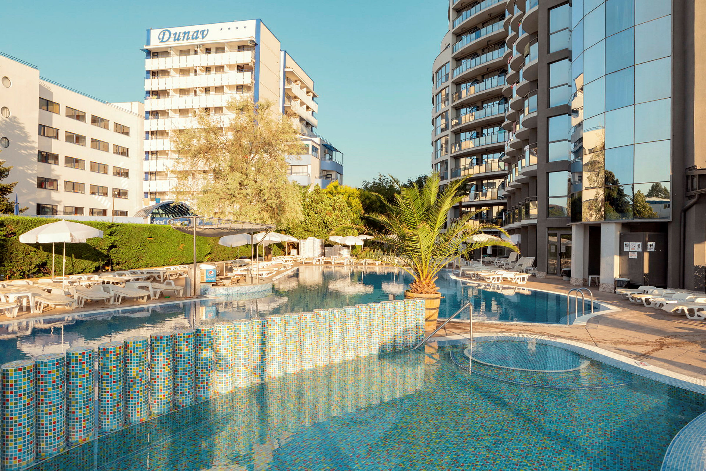 Bilde av hotellområde med utendørsbasseng, omgitt av solstoler og parasoller. Bakgrunn med bygninger og vegetasjon. (AI-generert tekst)
