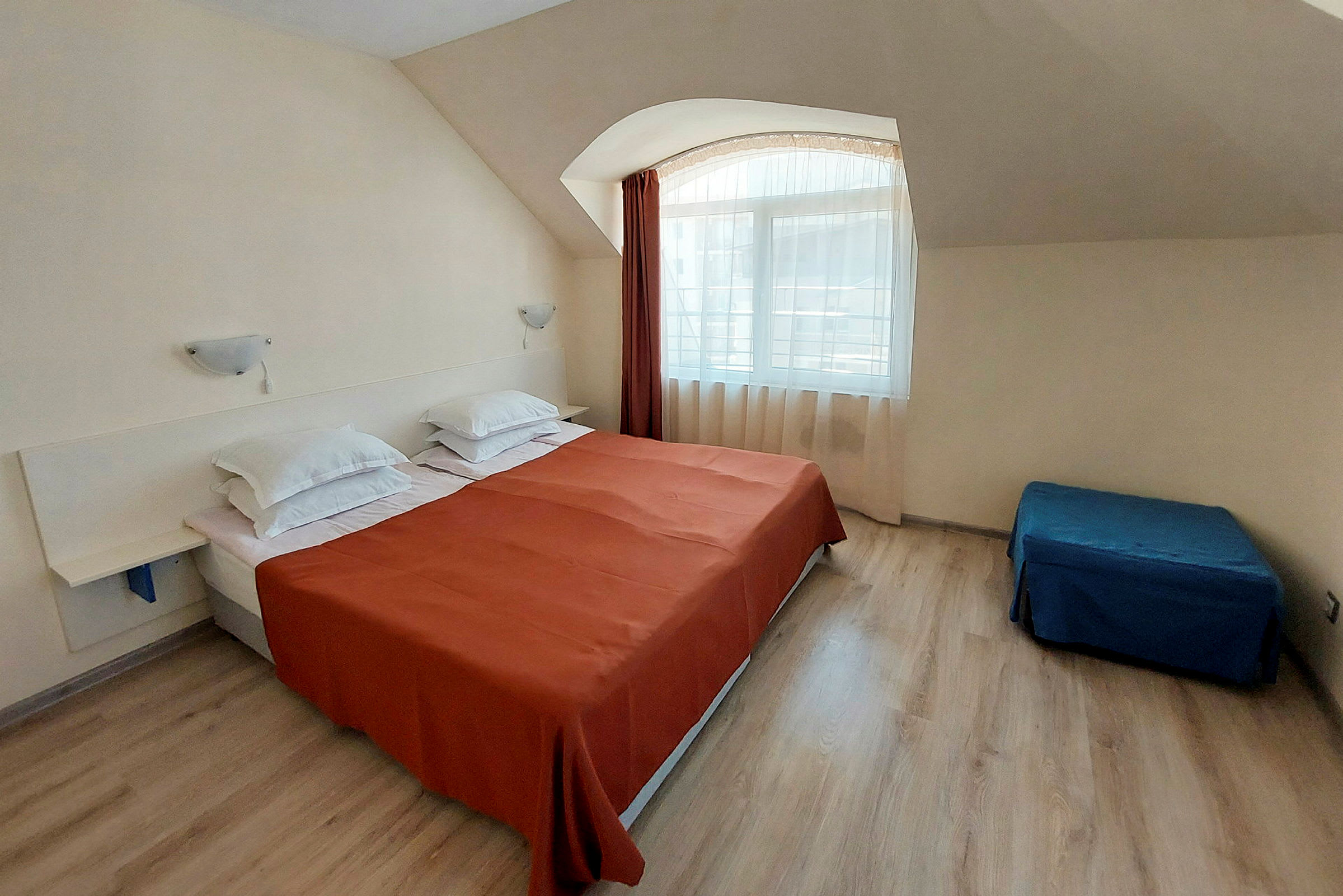Et hotellrom med en dobbeltseng kledd med oransje sengeteppe og hvite puter, en blå sittepuff og et vindu med lyse gardiner. (AI-generert tekst)