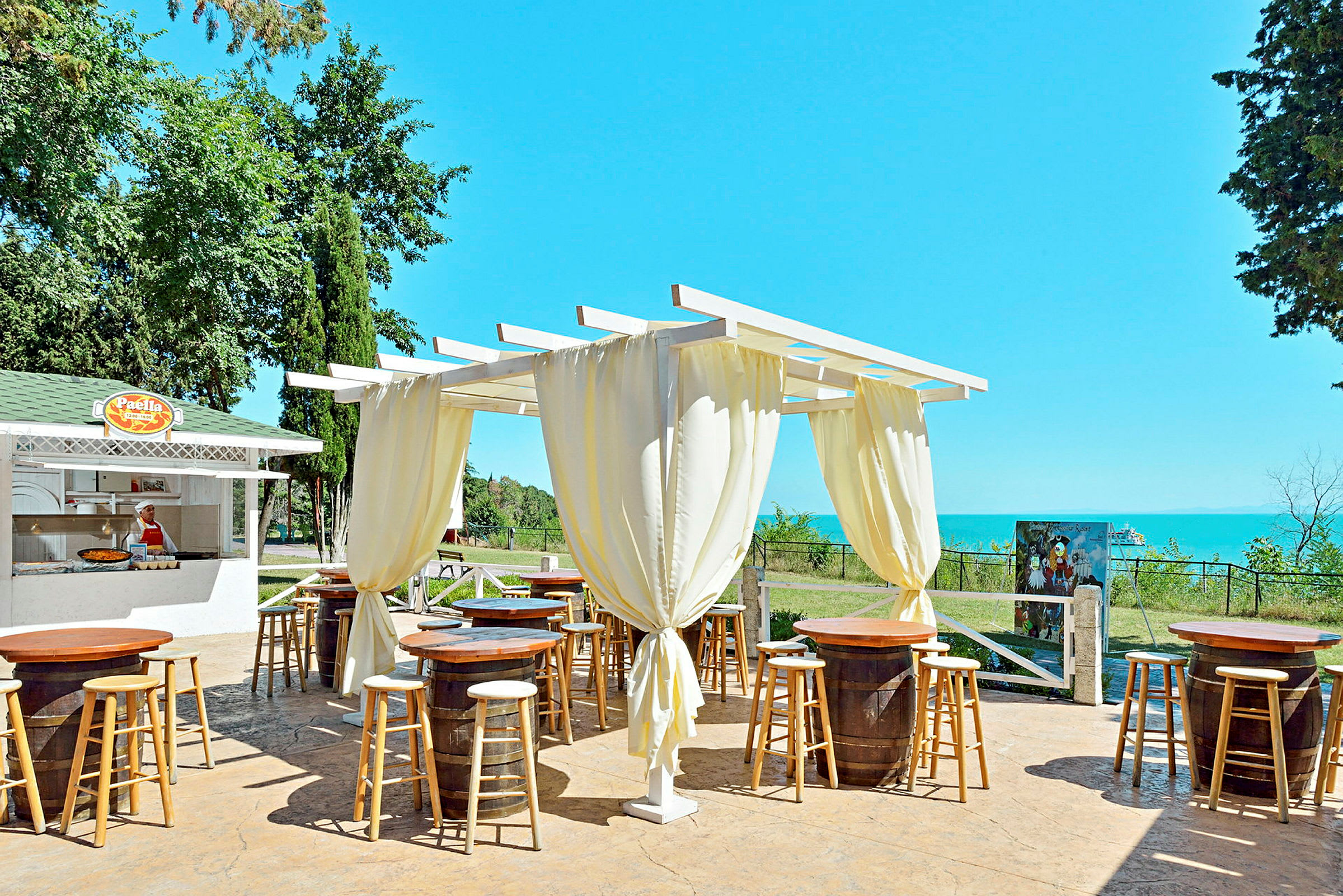 En utendørs restaurant med pergola, oppstilte trebord og stoler samt en matservering i bakgrunnen. (AI-generert tekst)