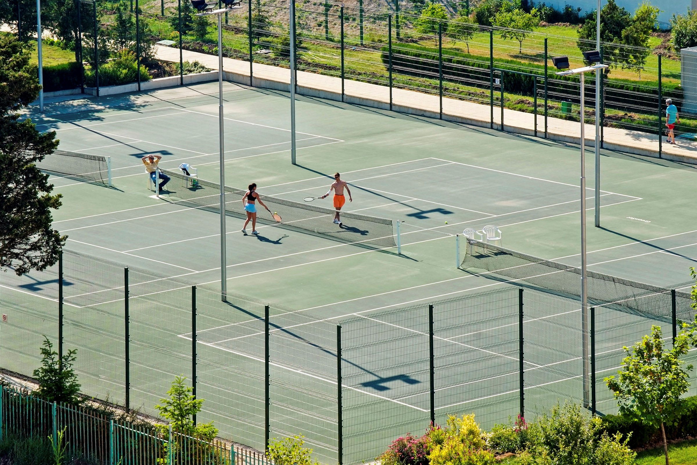 To personer spiller tennis på en utendørsbane omgitt av grøntområder. (AI-generert tekst)