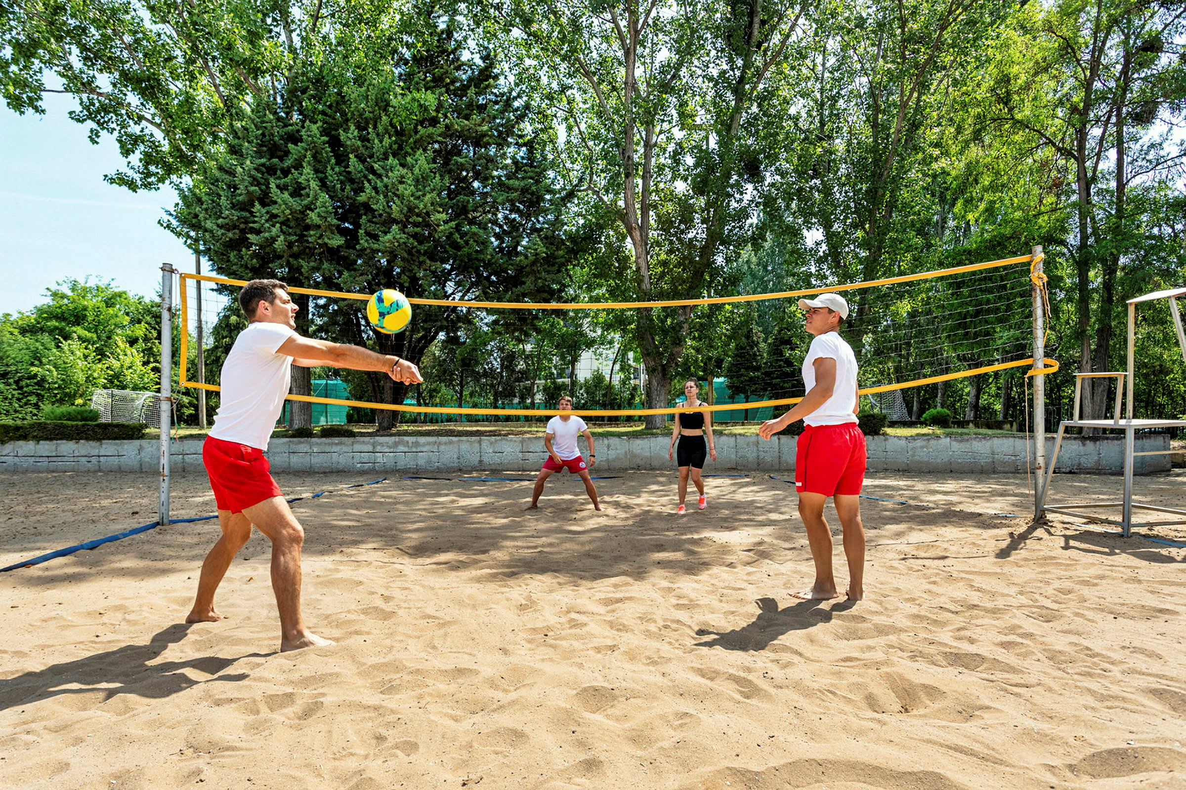 Personer spiller volleyball på en sandbane utendørs omgitt av grønne trær. (AI-generert tekst)