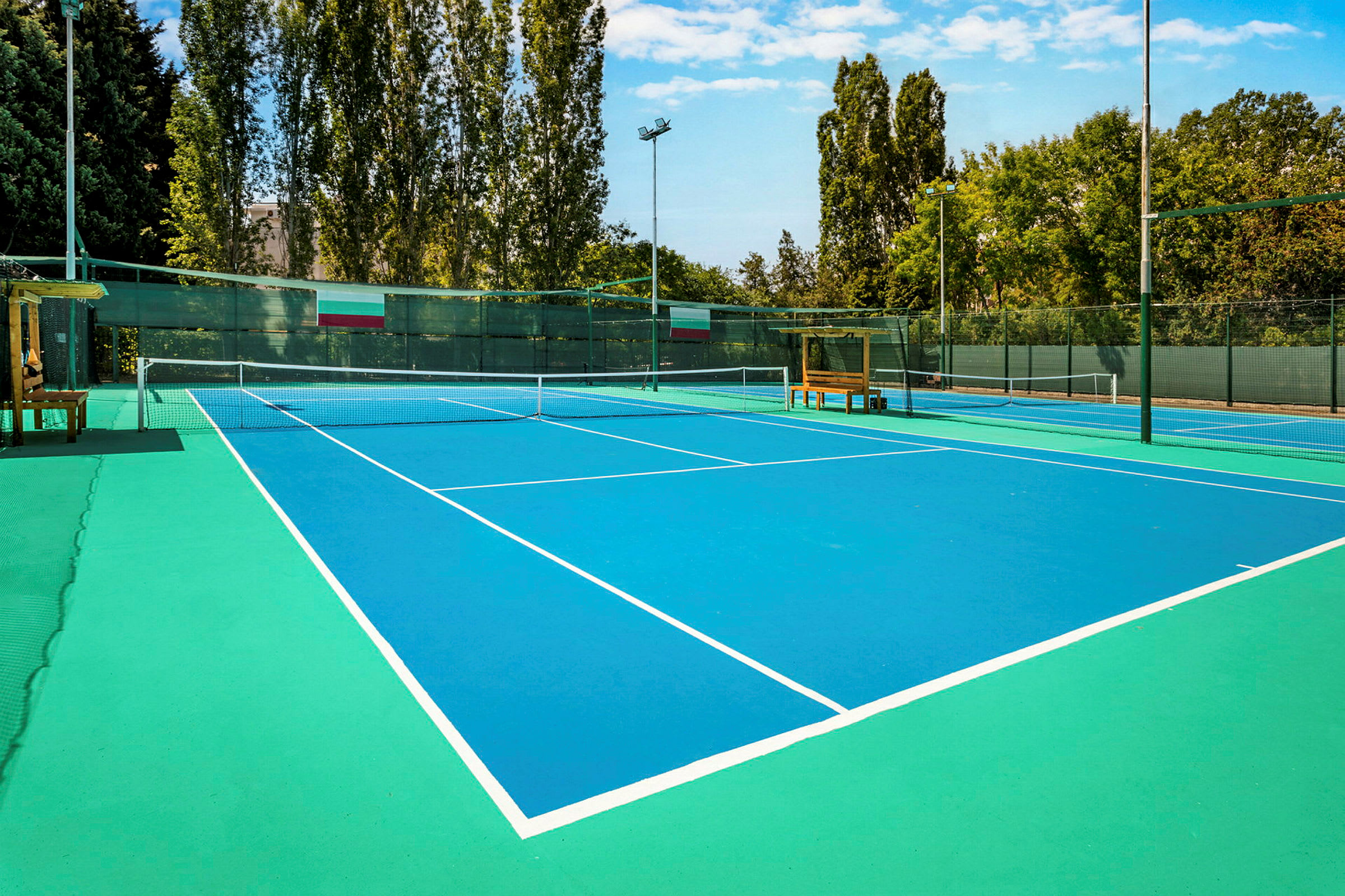 Bilde av en tennisbane med blå overflate og hvite linjer, omgitt av trær og gjerde. (AI-generert tekst)