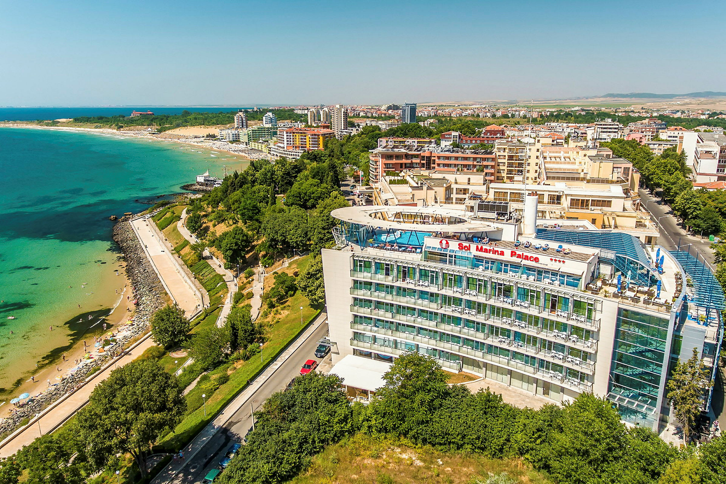 Luftfoto av et hotell med teksten "Sol Marina Palace", plassert nær en strand og kystlinje med vegetasjon og bygninger i bakgrunnen. (AI-generert tekst)