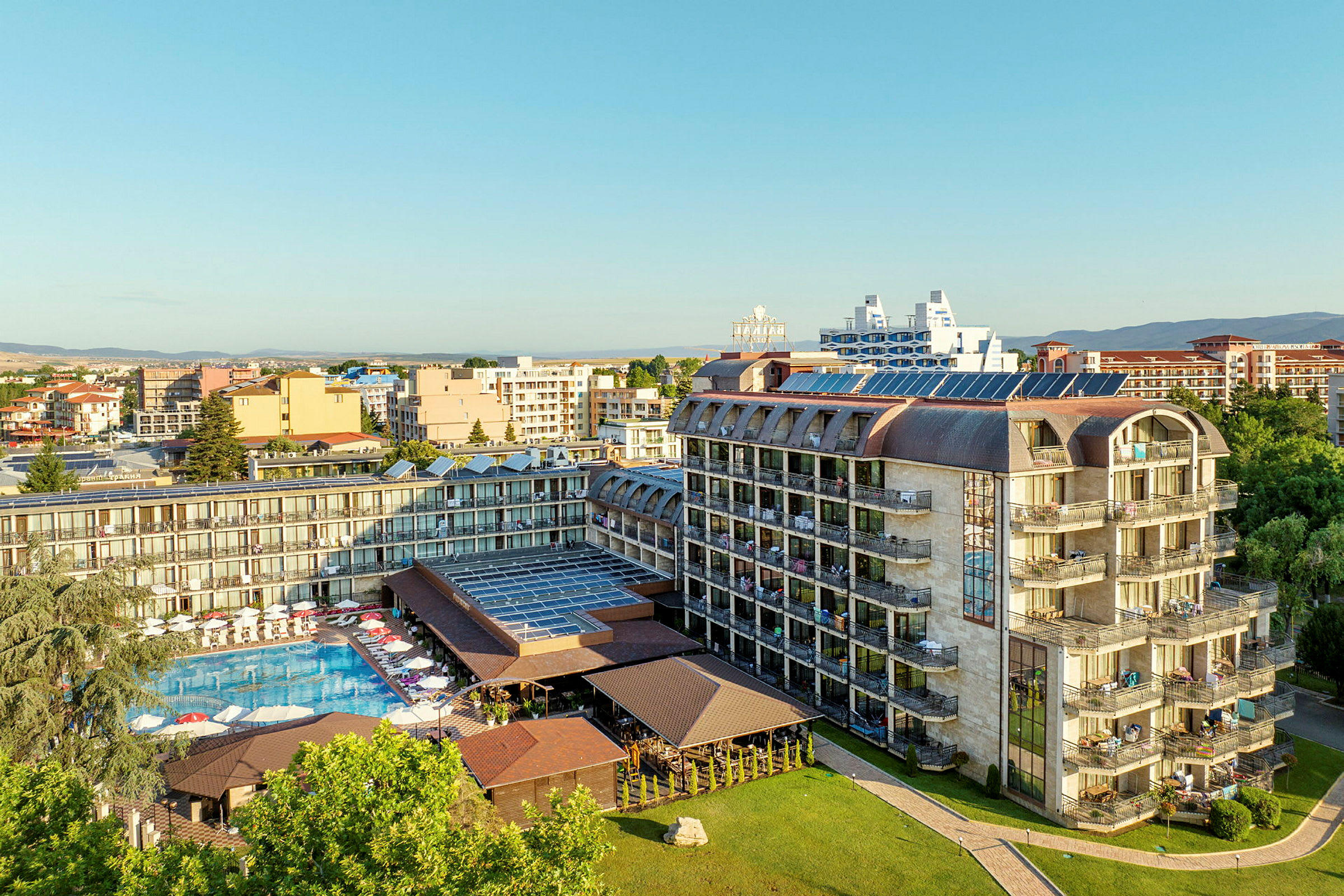 Luftfoto av et hotell med svømmebasseng og solsenger, omgitt av andre bygninger og grøntområder. (AI-generert tekst)