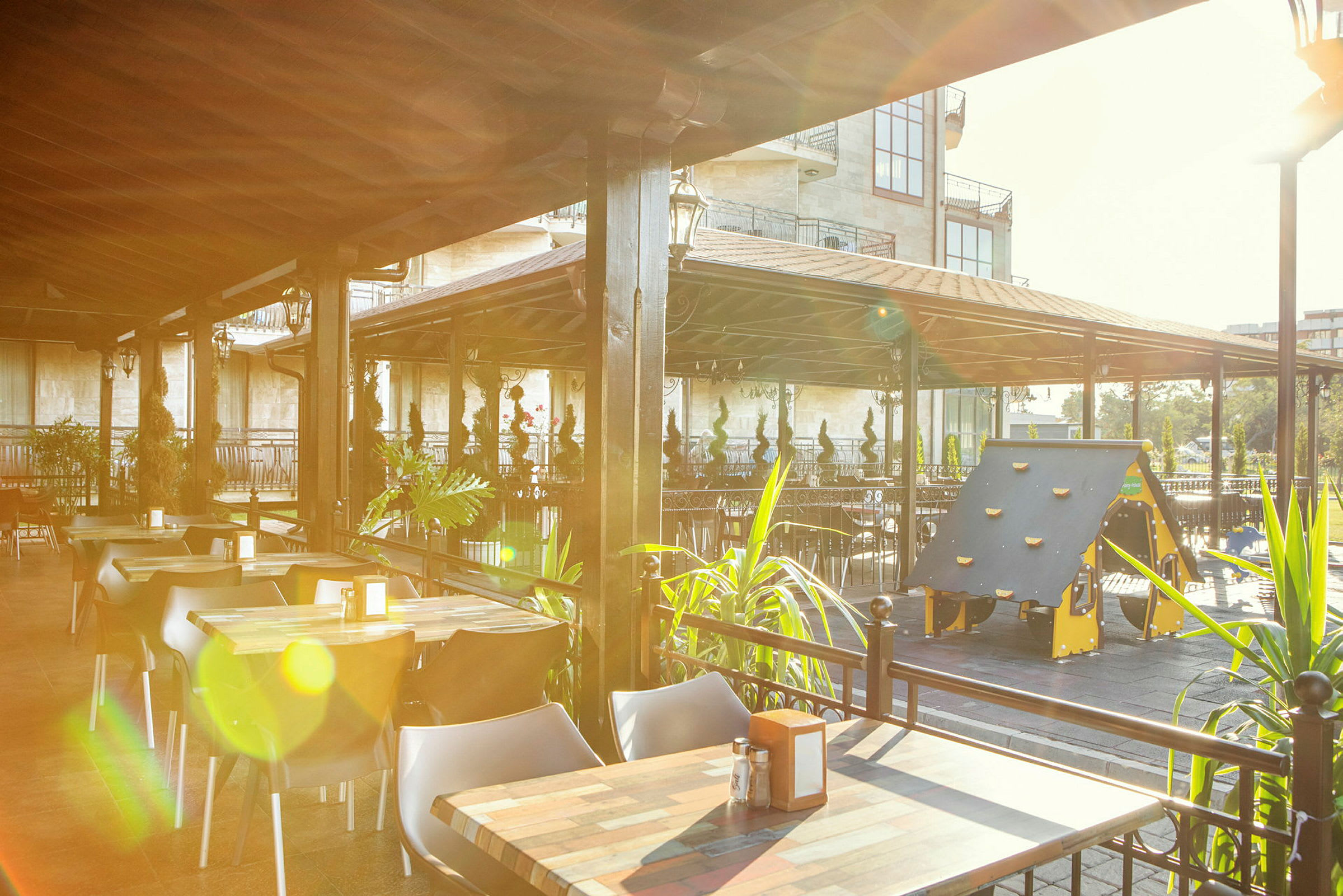 En utendørsrestaurant med bord, stoler og en lekeplass for barn under et tak. (AI-generert tekst)