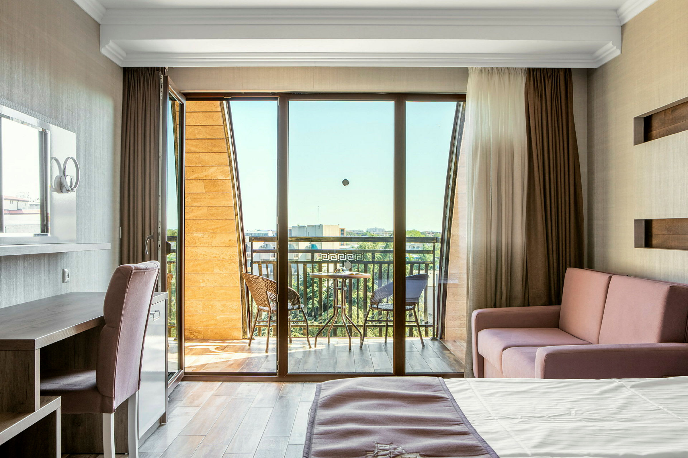 Et lyst hotellrom med balkong, møblert med seng, sofa og skrivebord. (AI-generert tekst)