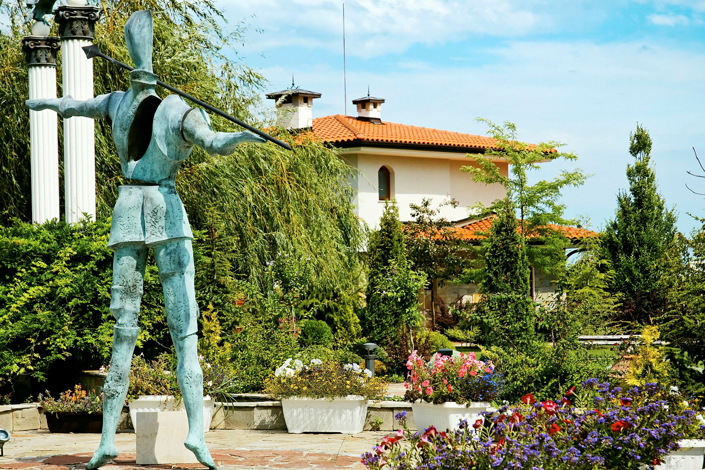 En statue i en hage omgitt av blomster og grønn buskvegetasjon. I bakgrunnen sees et hus med oransjet tak. (AI-generert tekst)