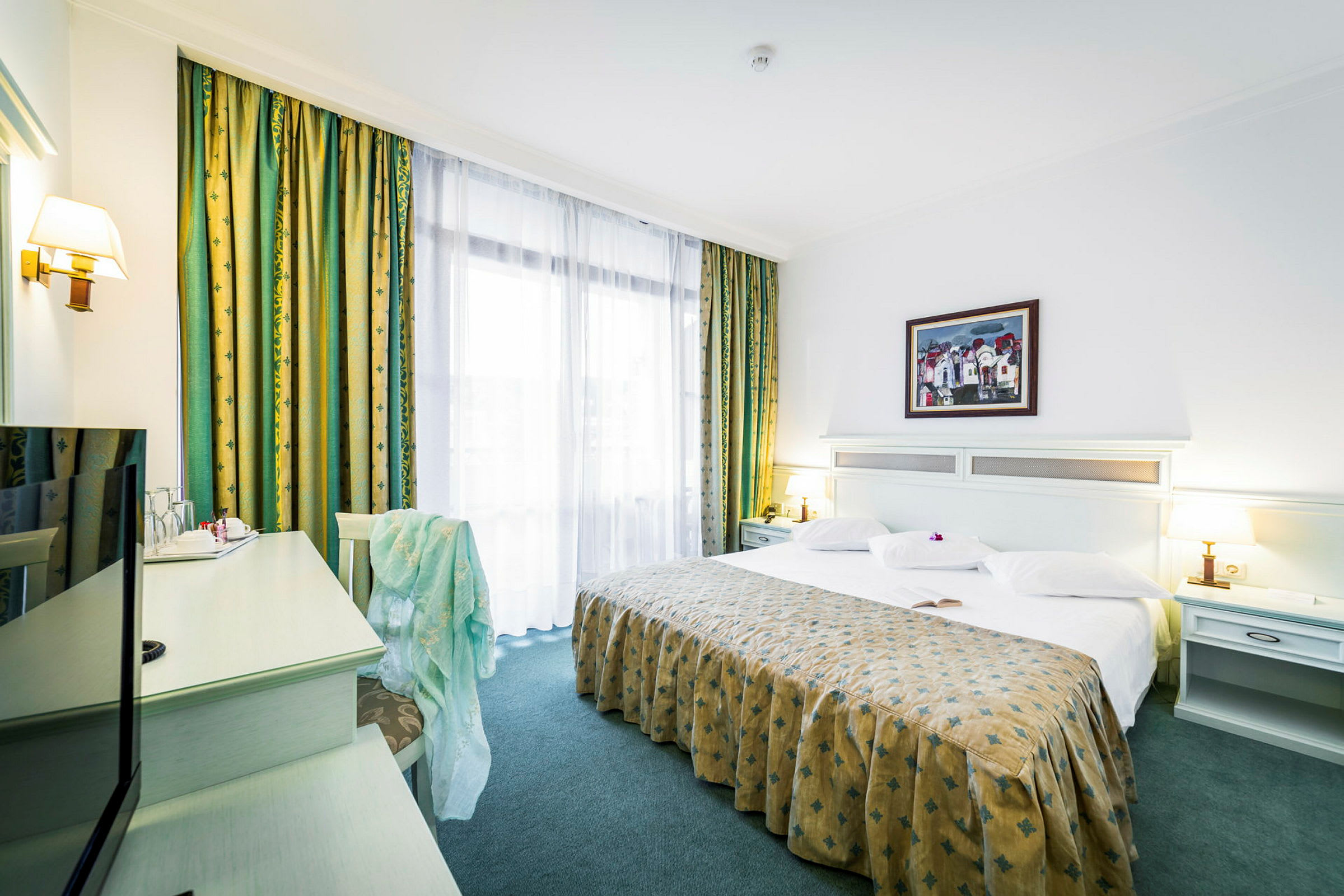 Et hotellrom med en stor seng, hvitt sengetøy og et mønstret sengeteppe. Det finnes bordlamper ved siden av sengen og et skrivebord med en stol i hjørnet. (AI-generert tekst)