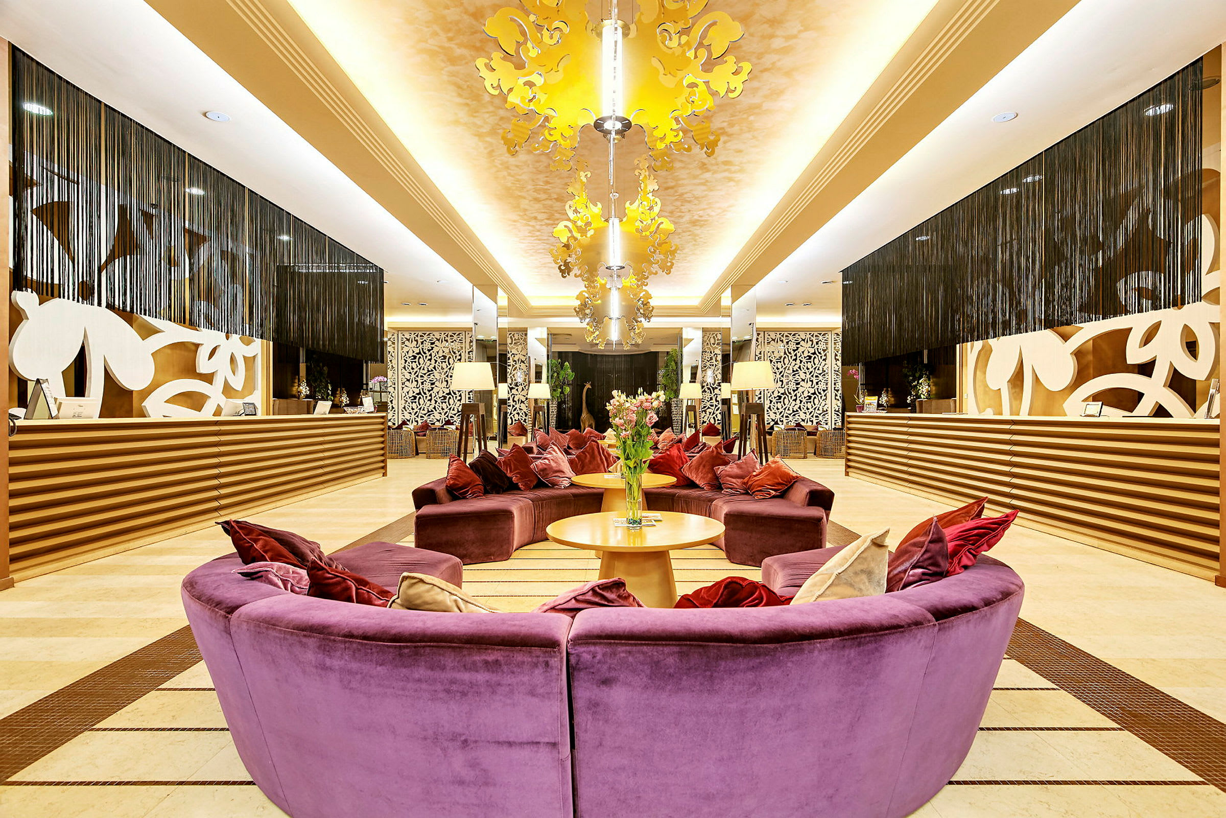 Resepsjonen på et hotell med elegant interiør. Fargerike sofaer i lilla med puter er plassert i midten, med to bord og blomster sentrert. Bak resepsjonen finnes dekorative mønstre og hengende gardiner. (AI-generert tekst)