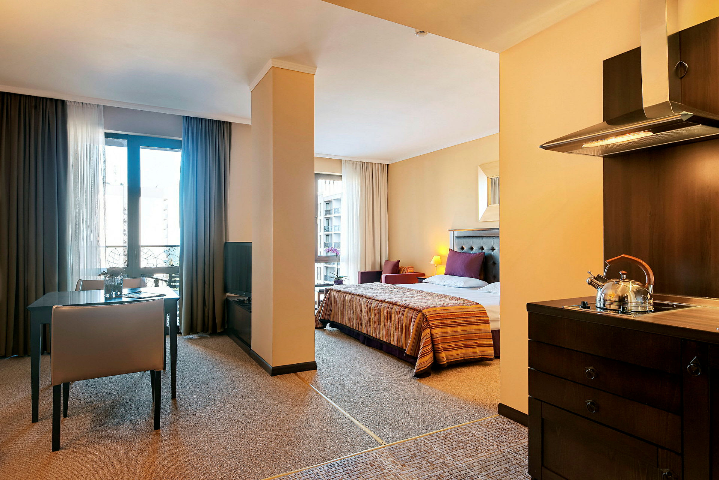 Interiør av et hotellrom med soveplass, spisebord og kjøkken. Sengen er oppredd og kjøkkenet har en tekanne på komfyren. (AI-generert tekst)