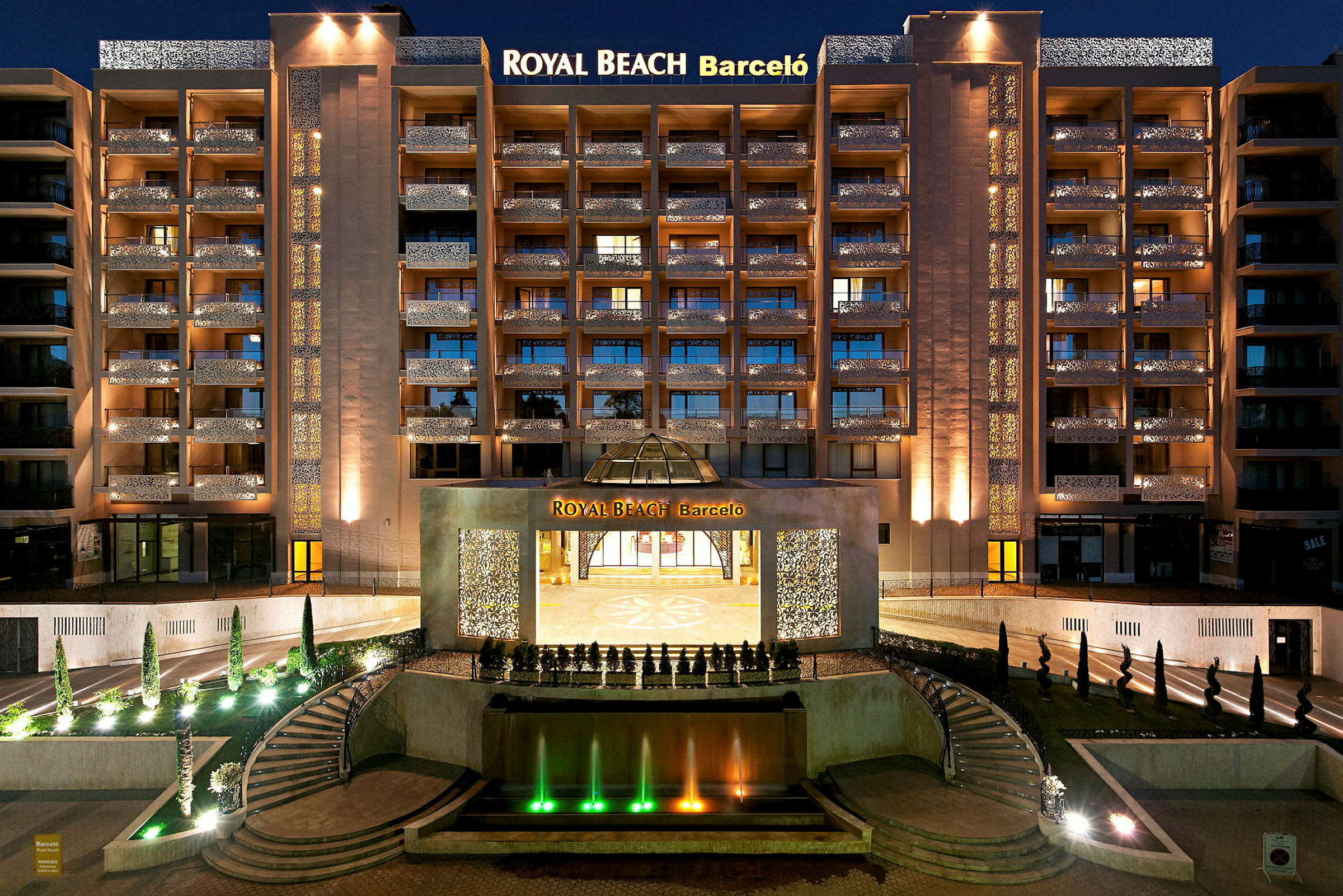 Et nattbilde av et opplyst hotell med navnet Royal Beach Barceló, med en fontene og trapper i forgrunnen. (AI-generert tekst)
