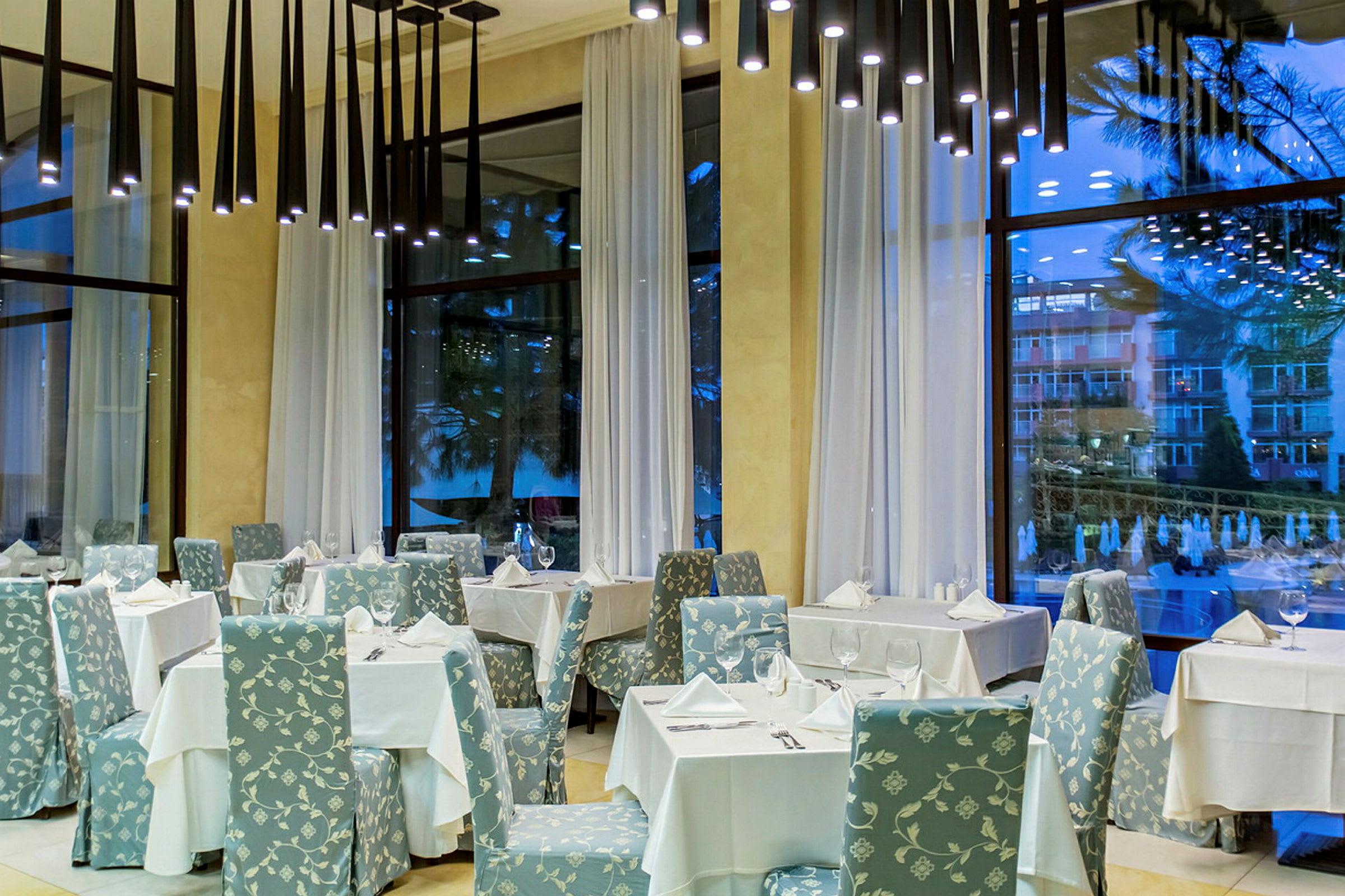 En elegant restaurant med bord dekket med hvite duker og stoler med blomstrete mønster, høye vinduer med hvite gardiner. (AI-generert tekst)