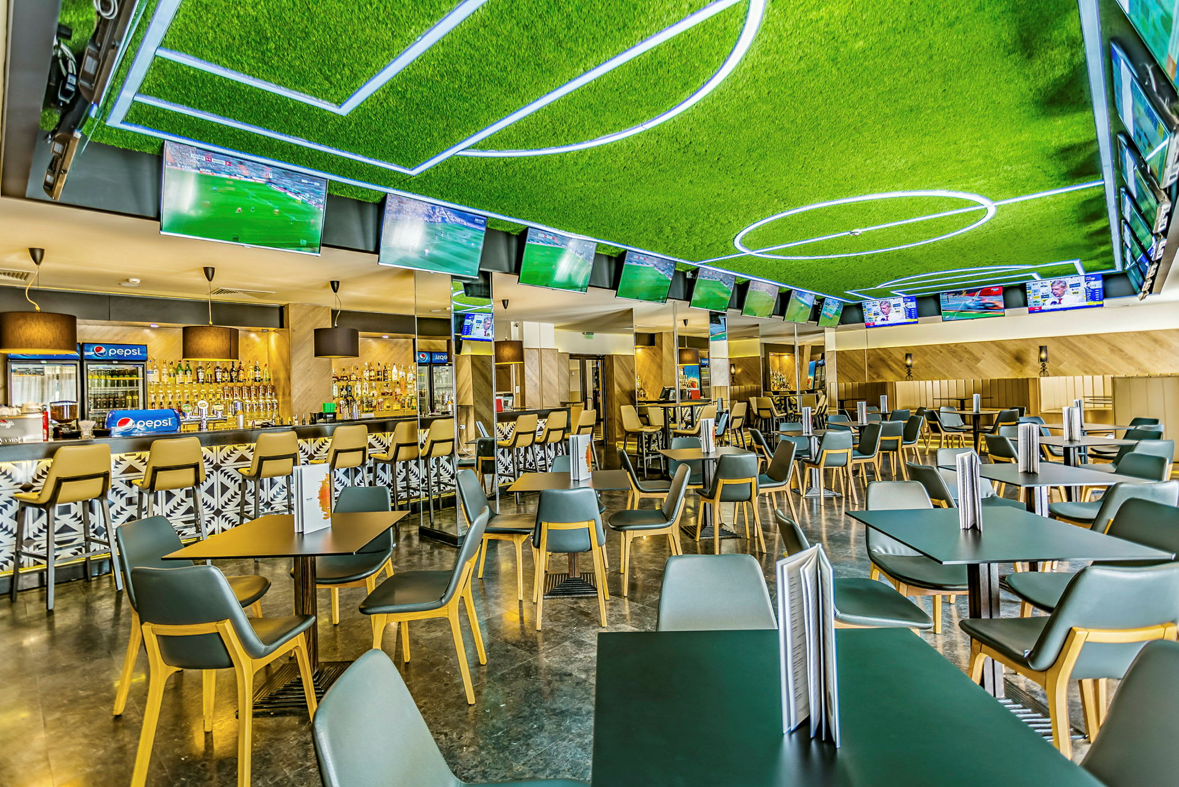 Et rom med flere bord og stoler, en bar i bakgrunnen og TV-skjermer som viser fotballkamper. (AI-generert tekst)