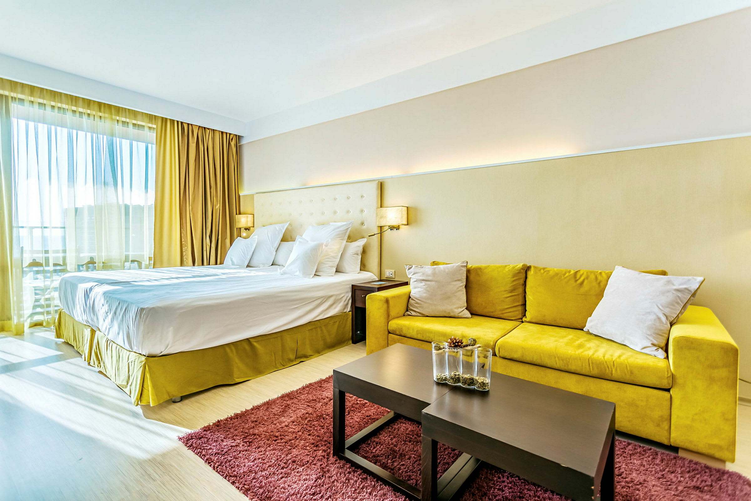 Et hotellrom med en stor dobbeltseng, gul sofa, et bord med dekorativ vase og et rødt teppe. (AI-generert tekst)