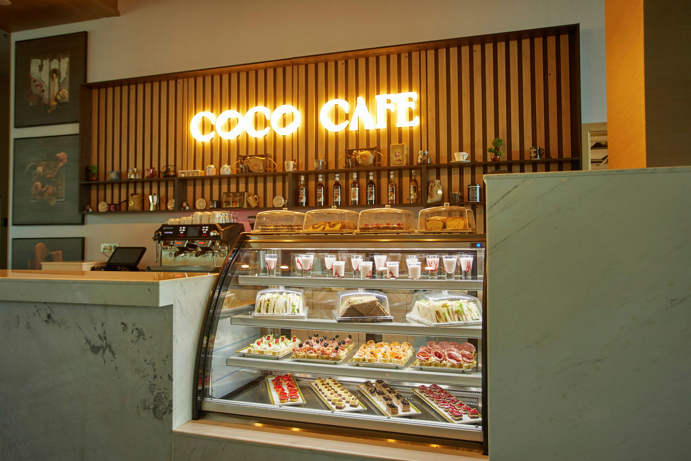 En kafédisk med et opplyst skilt som sier 'Coco Cafe' og et utvalg bakverk og drikker bak glass. (AI-generert tekst)