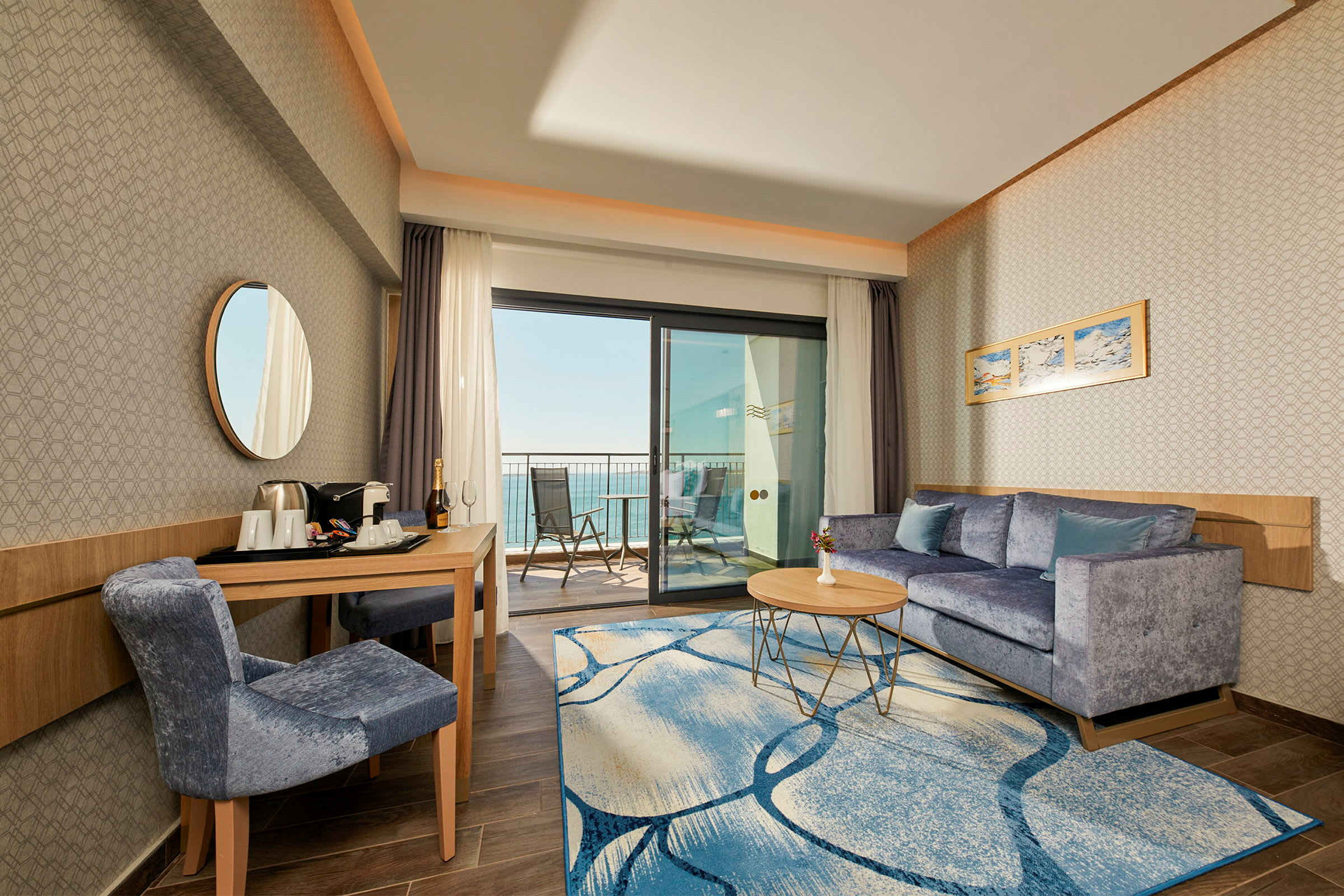 Hotellrom med sittegruppe, bord og stol samt balkong med utsikt mot havet. (AI-generert tekst)