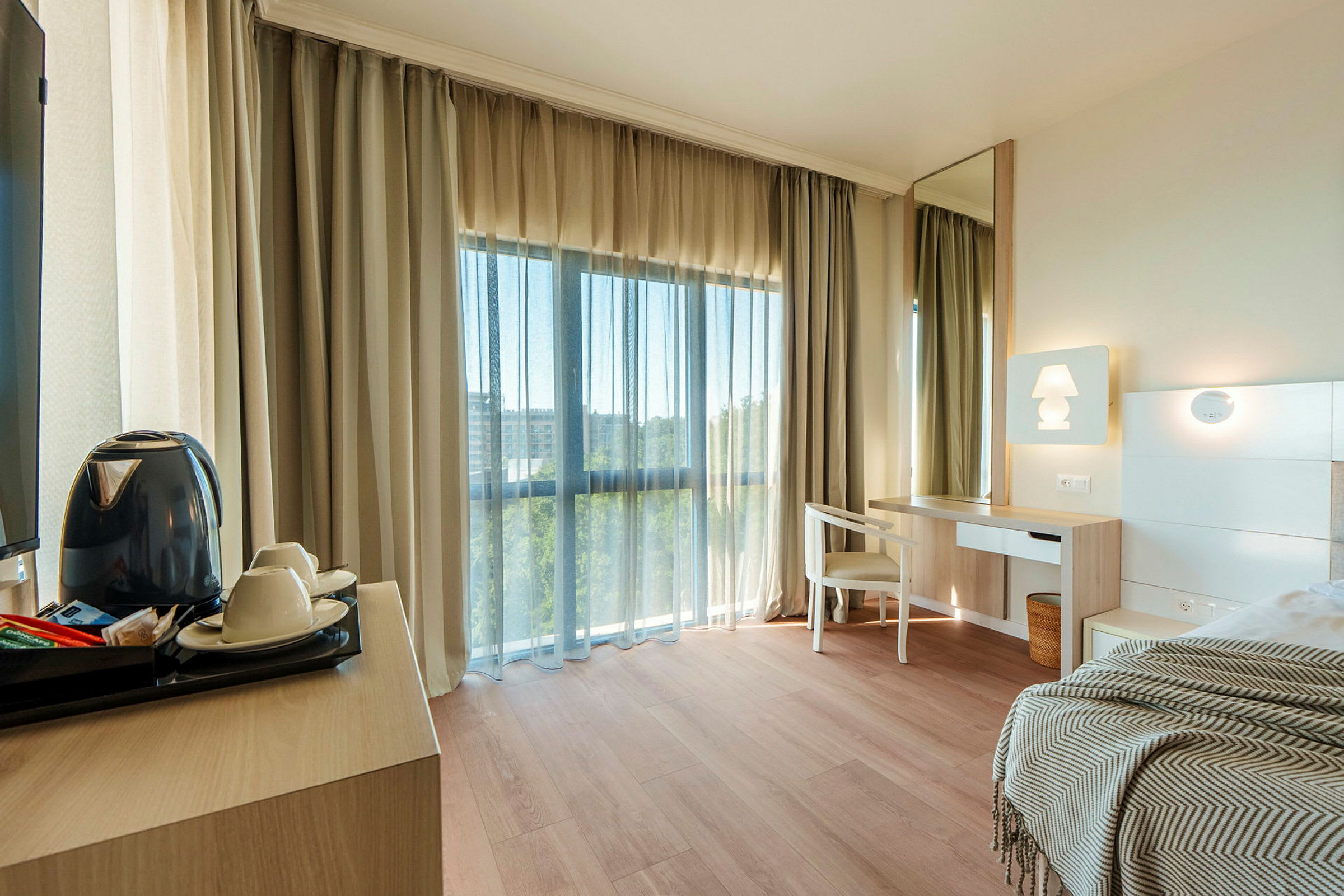 Et lyst hotellrom med tregulv, et skrivebord med stol og speil, samt en seng med et teppe. Et stort vindu med gardiner slipper inn dagslys. (AI-generert tekst)