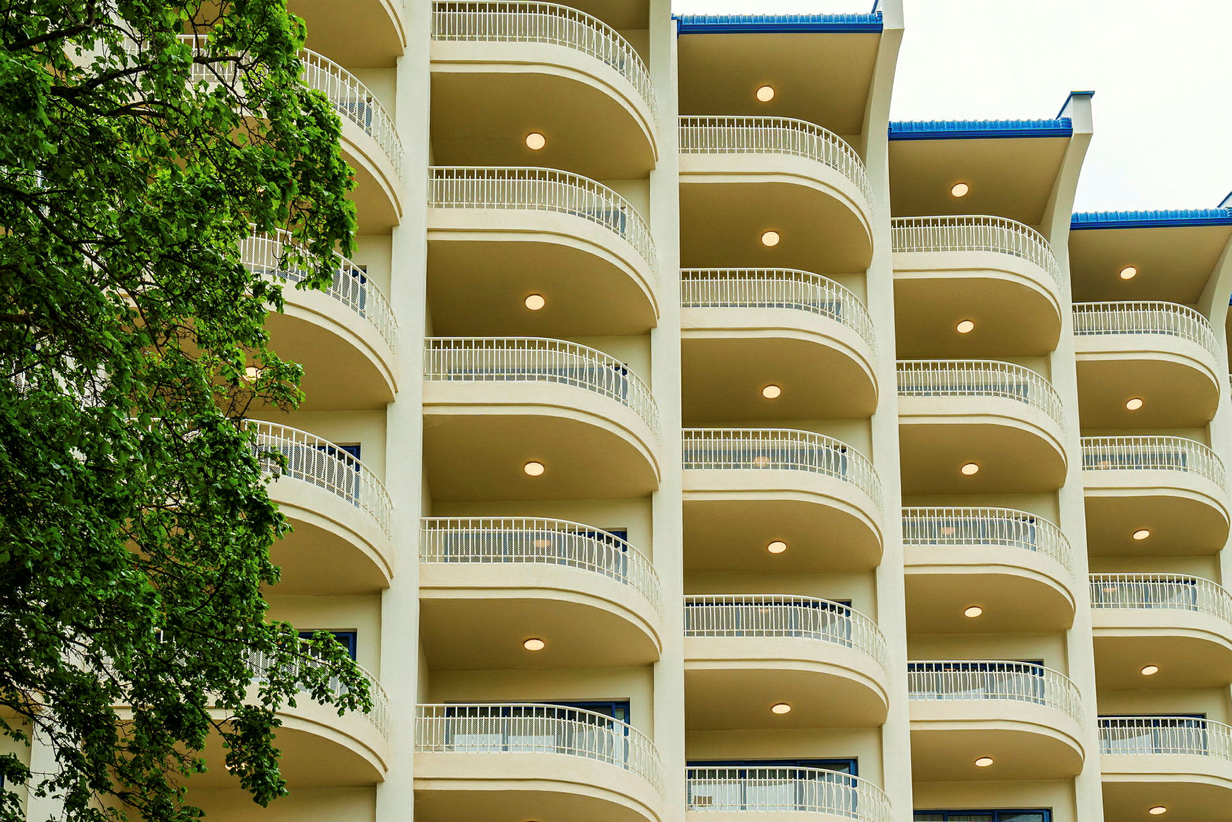 Fasaden til et hotell, med flere etasjer og balkonger. Et tre er til venstre i bildet. (AI-generert tekst)