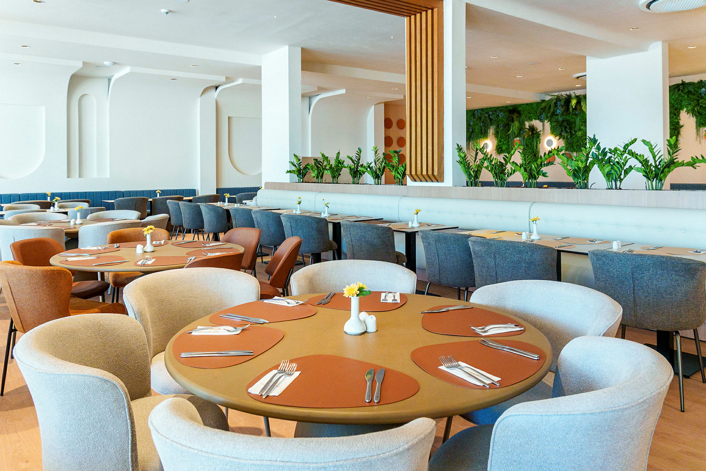 Et restaurantinteriør med runde og rektangulære bord, elegante stoler og grønne planter som dekor. (AI-generert tekst)