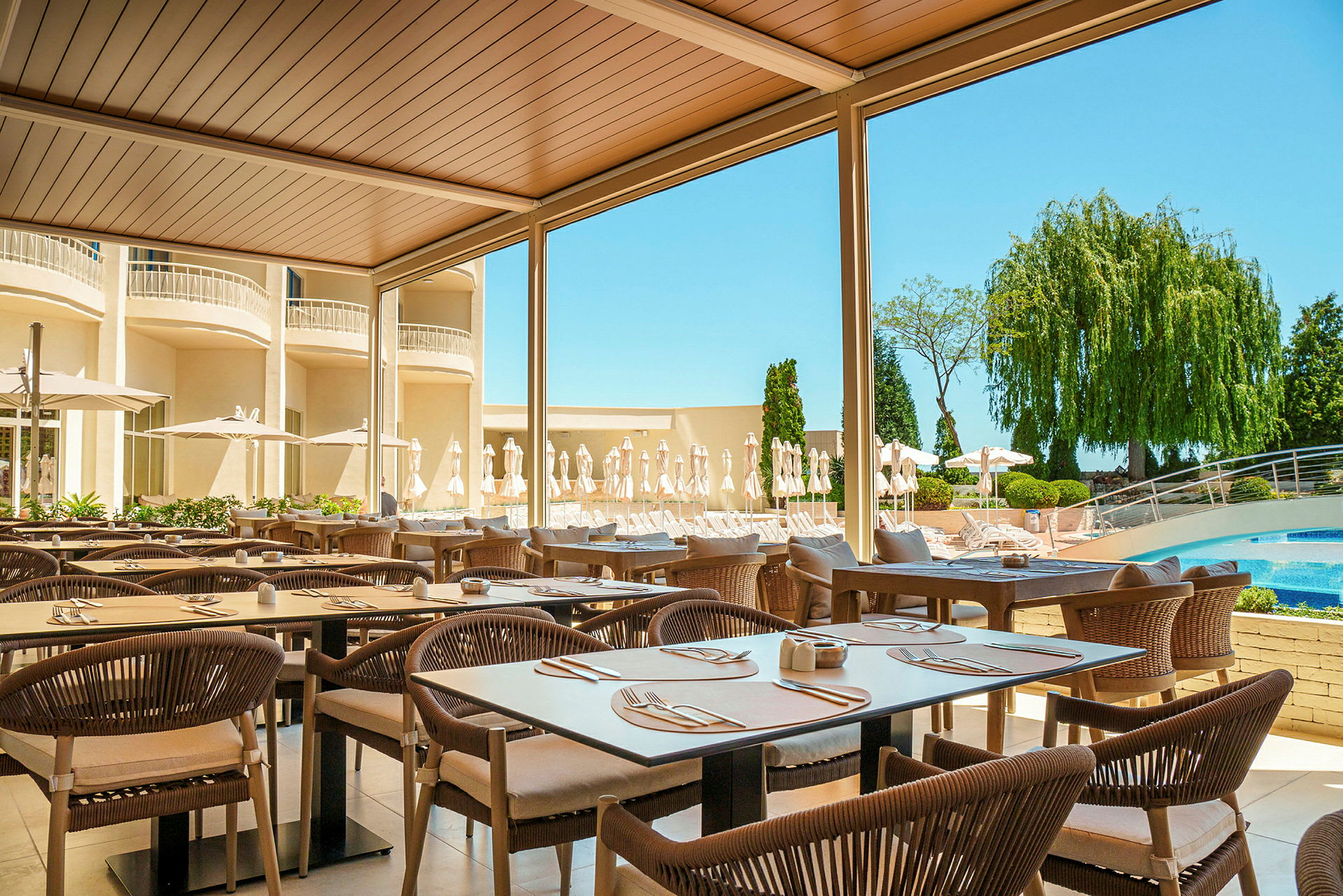 Utendørsrestaurant med bord og stoler under tak, omgitt av grønt og et basseng i bakgrunnen. (AI-generert tekst)