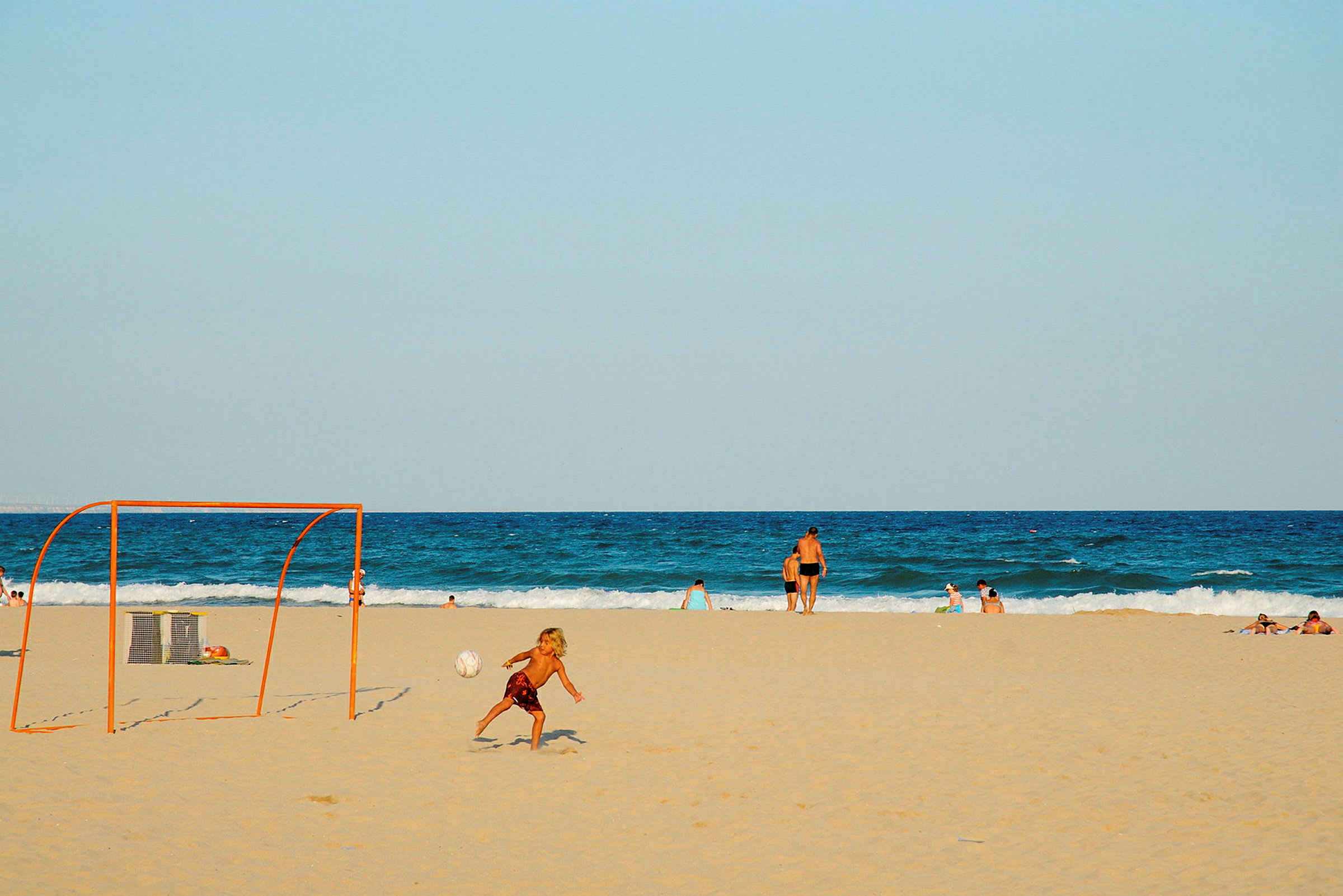En strand med mennesker som svømmer og et sandfotballmål. (AI-generert tekst)