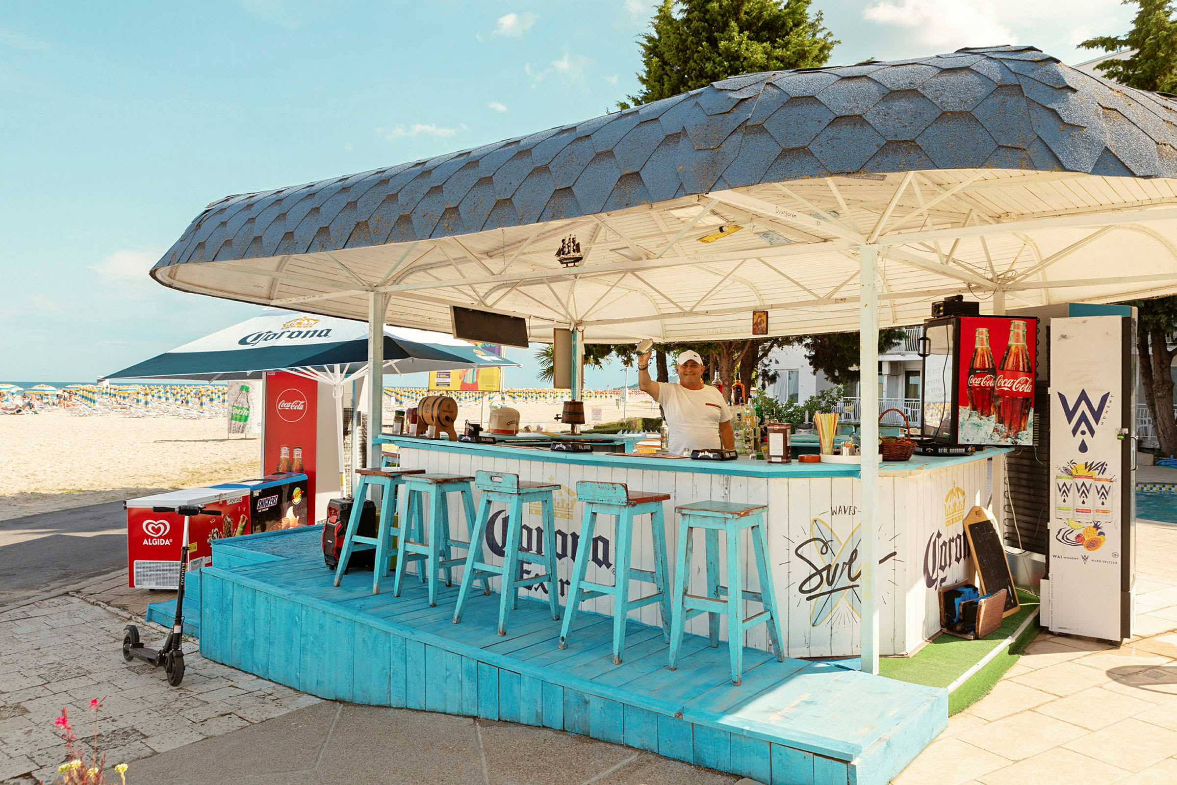 En strandbar med blå stoler og tak, plassert nær sandstranden. (AI-generert tekst)